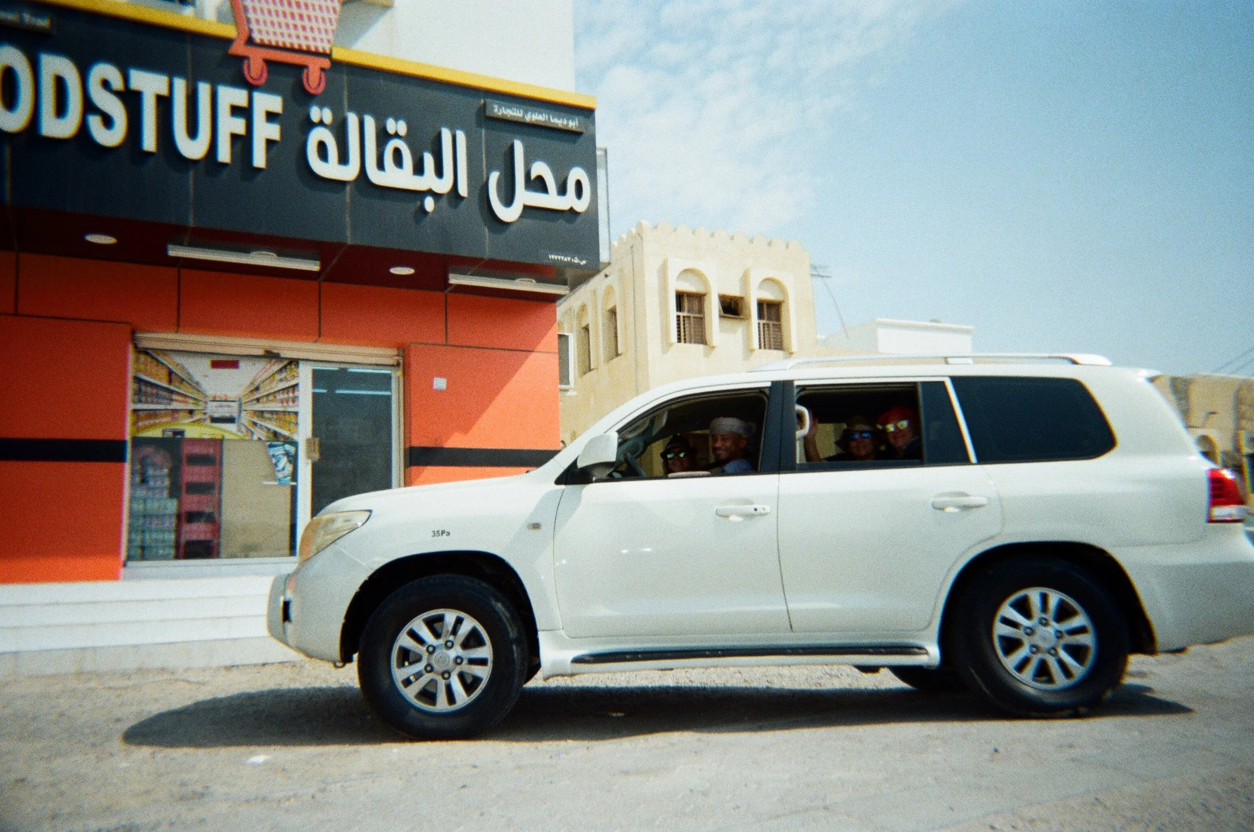 Auto bianca con quattro persone al suo interno, parcheggiata davanti a un negozio con cartelli in arabo e inglese, in un'area urbana con edifici di stile mediorientale.