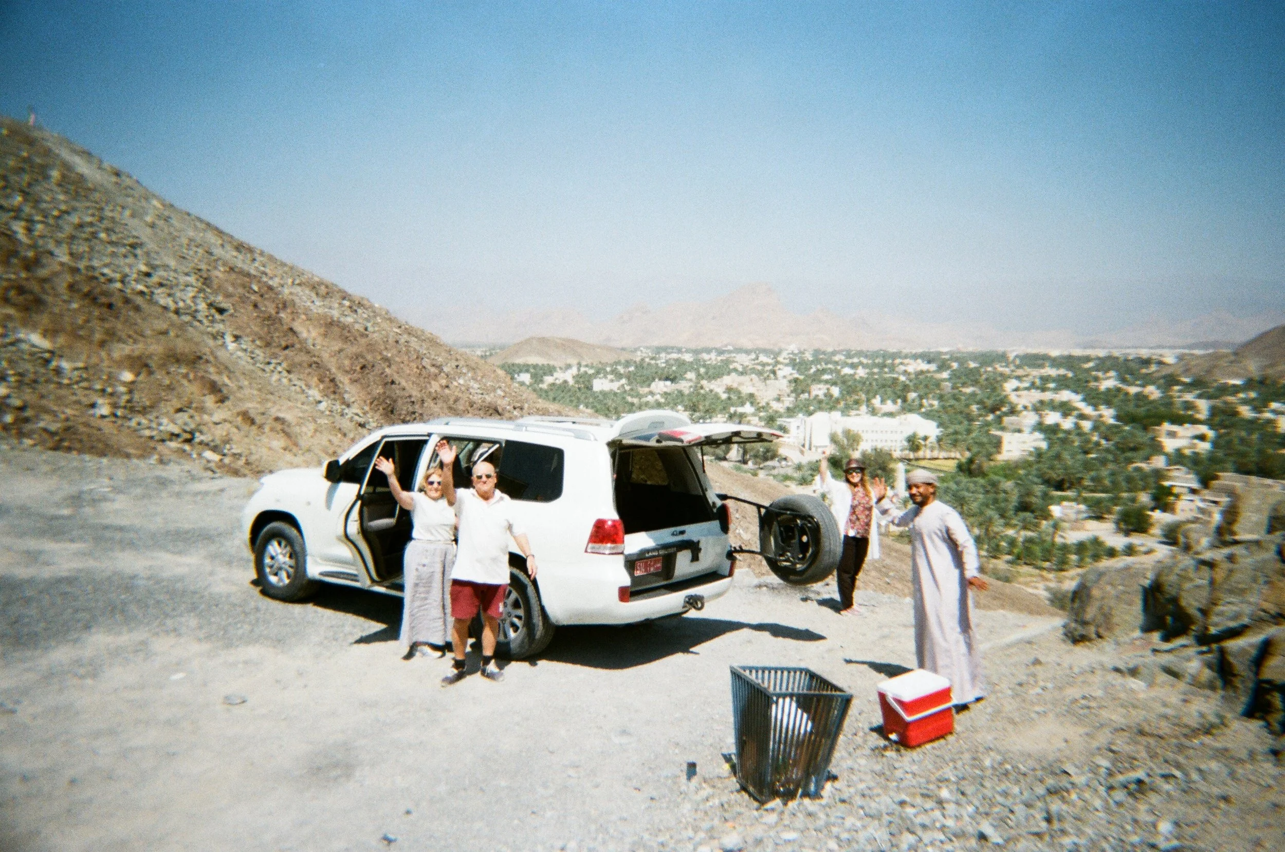 Gruppo di persone che si diverte accanto a un'auto in un paesaggio montano post desertico, con una città in lontananza sotto un cielo azzurro.