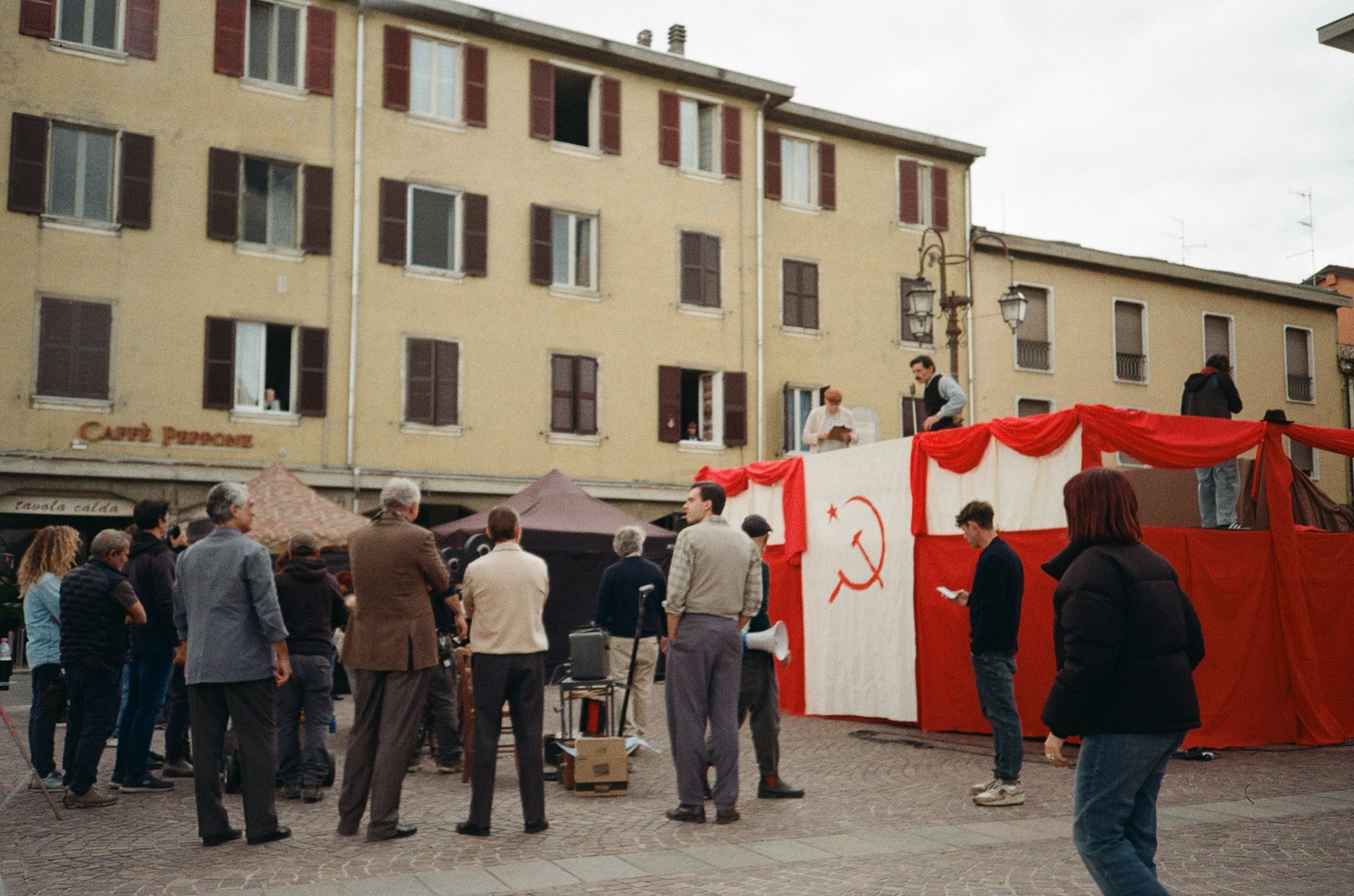 Una manifestazione o un evento pubblico con un palco decorato con una bandiera rossa e il simbolo del comunismo, con persone che si riuniscono in piazza.