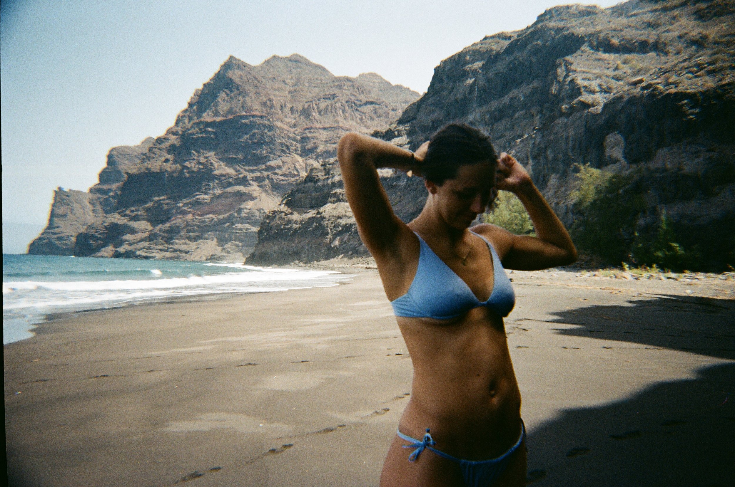 Donna in bikini blu sulla spiaggia con formazioni rocciose sullo sfondo.