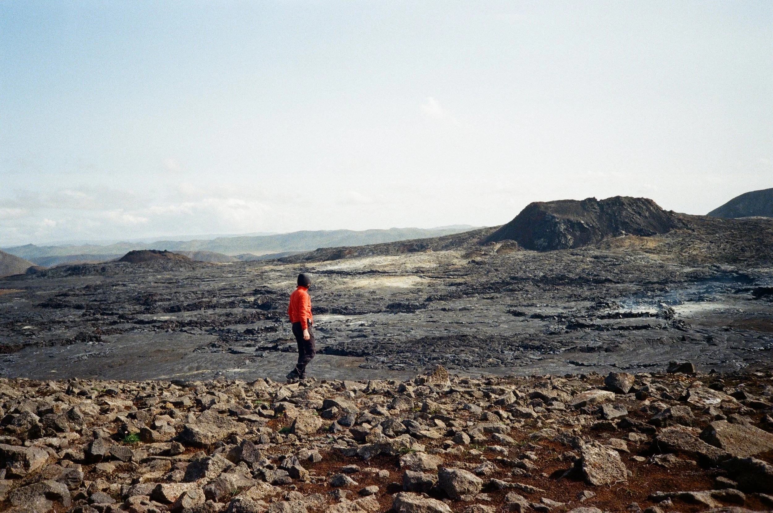 Un uomo con giacca rossa cammina su un paesaggio vulcanico di rocce nere e terra sconnessa, con montagne sullo sfondo e un cielo leggermente nuvoloso.