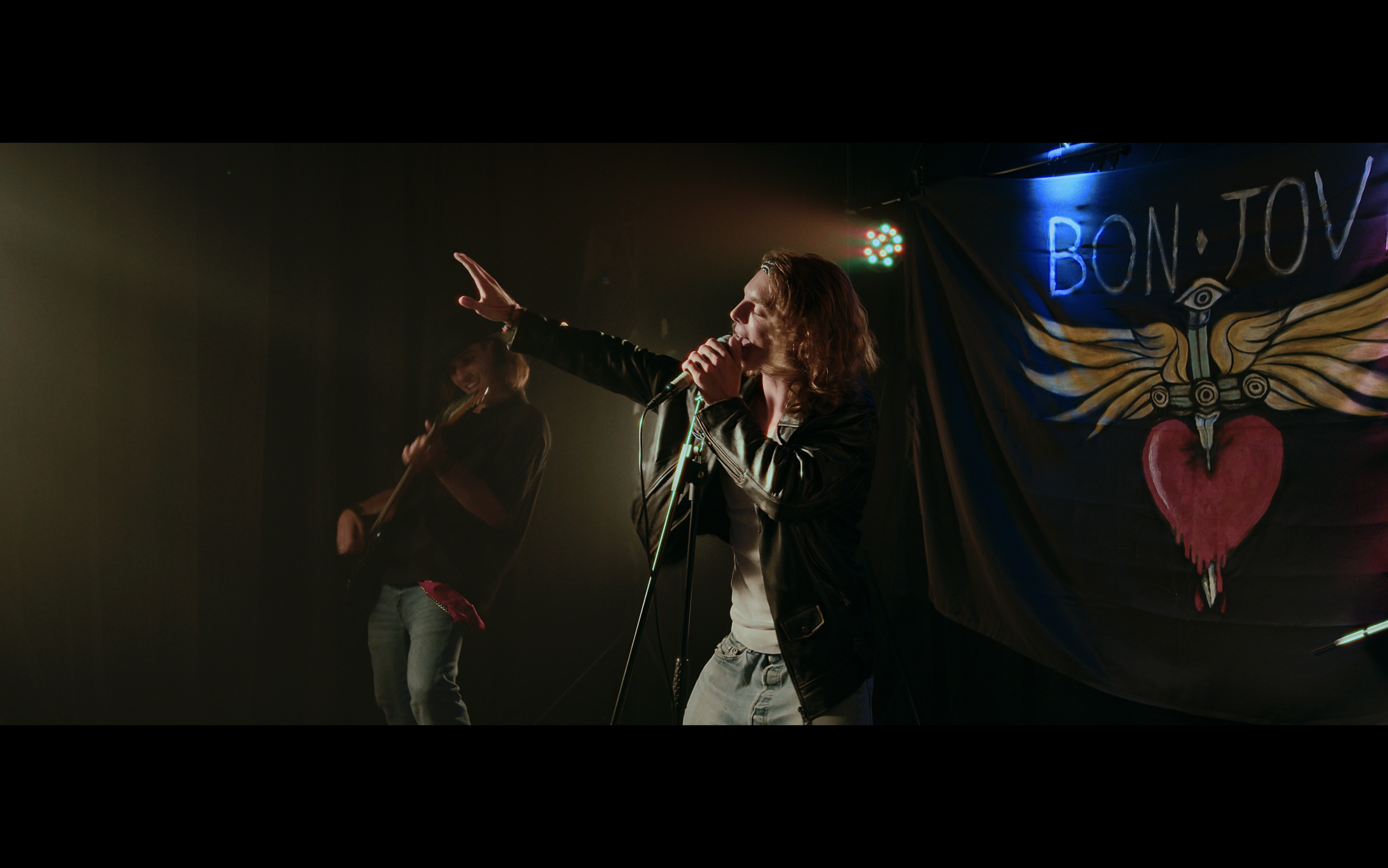 Un musicista con capelli ricci e grembiule di pelle canta sul palco con una voce potente, mentre un altro musicista in background suona la chitarra. Sullo sfondo, un cartellone con le parole 'BON JOVI' e un cuore con ali dipinte sopra.