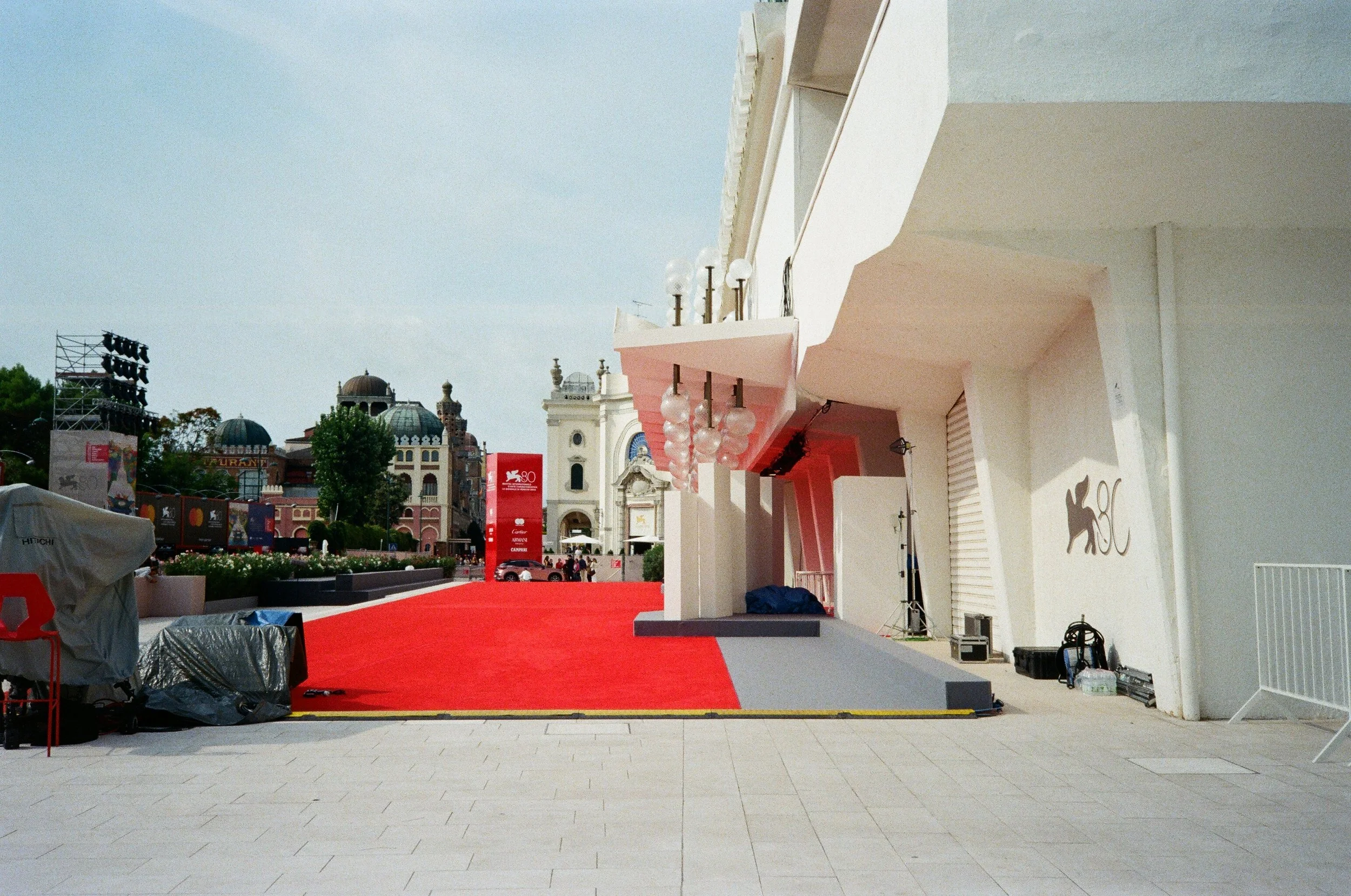 Area all'aperto con tappeto rosso, strutture moderne e edifici storici sullo sfondo. Attrezzature, sedie e luci pronte per un evento.