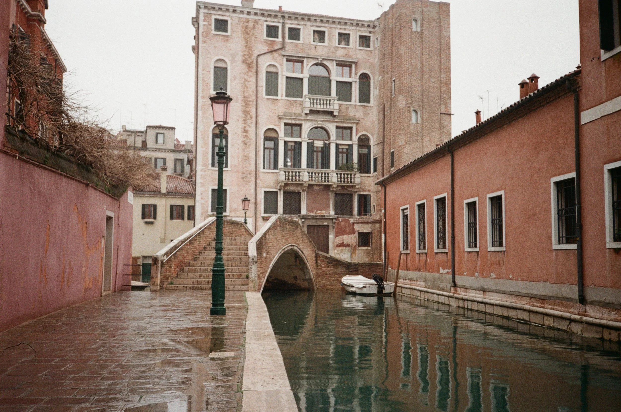 Veduta di un canale veneziano con un piccolo ponte, edifici antichi e una barca attraccata lungo il molo, in una giornata piovosa.