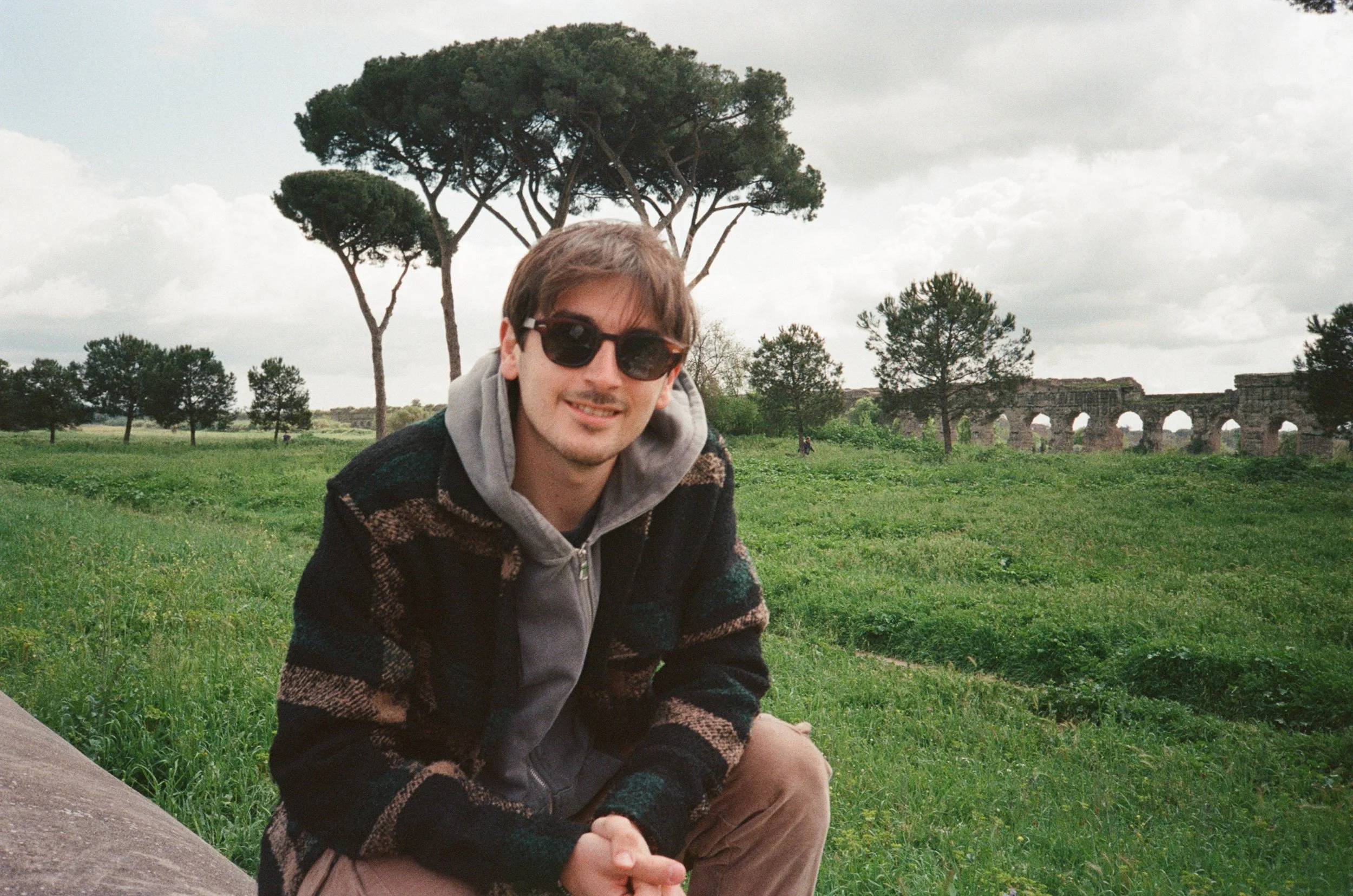 Persona sorridente con occhiali da sole in un parco verde con alberi e resti di un antico acquedotto in background.