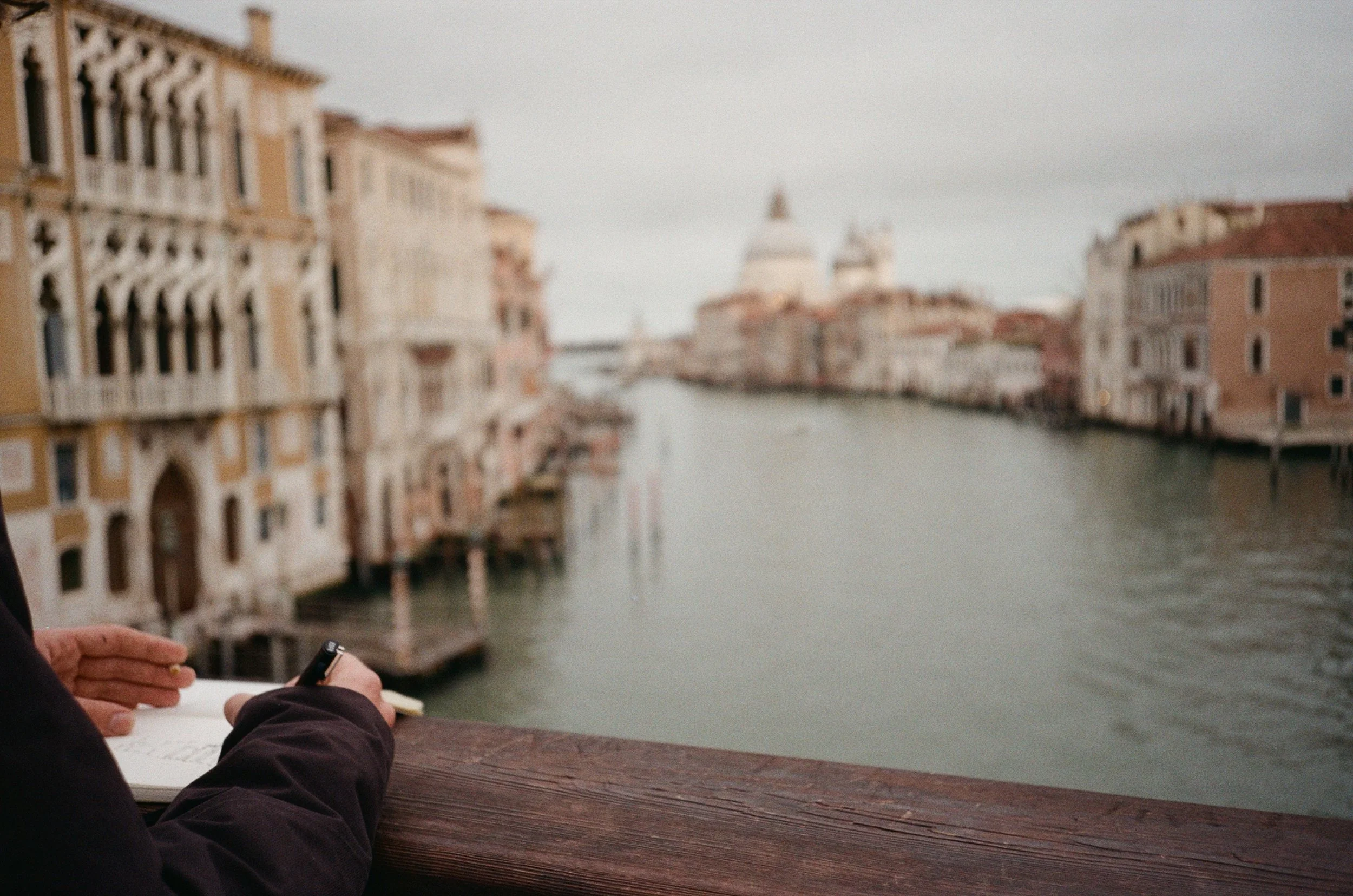 Persona che scrive in un quaderno con vista sul canale di Venezia e le sue edificazioni storiche.