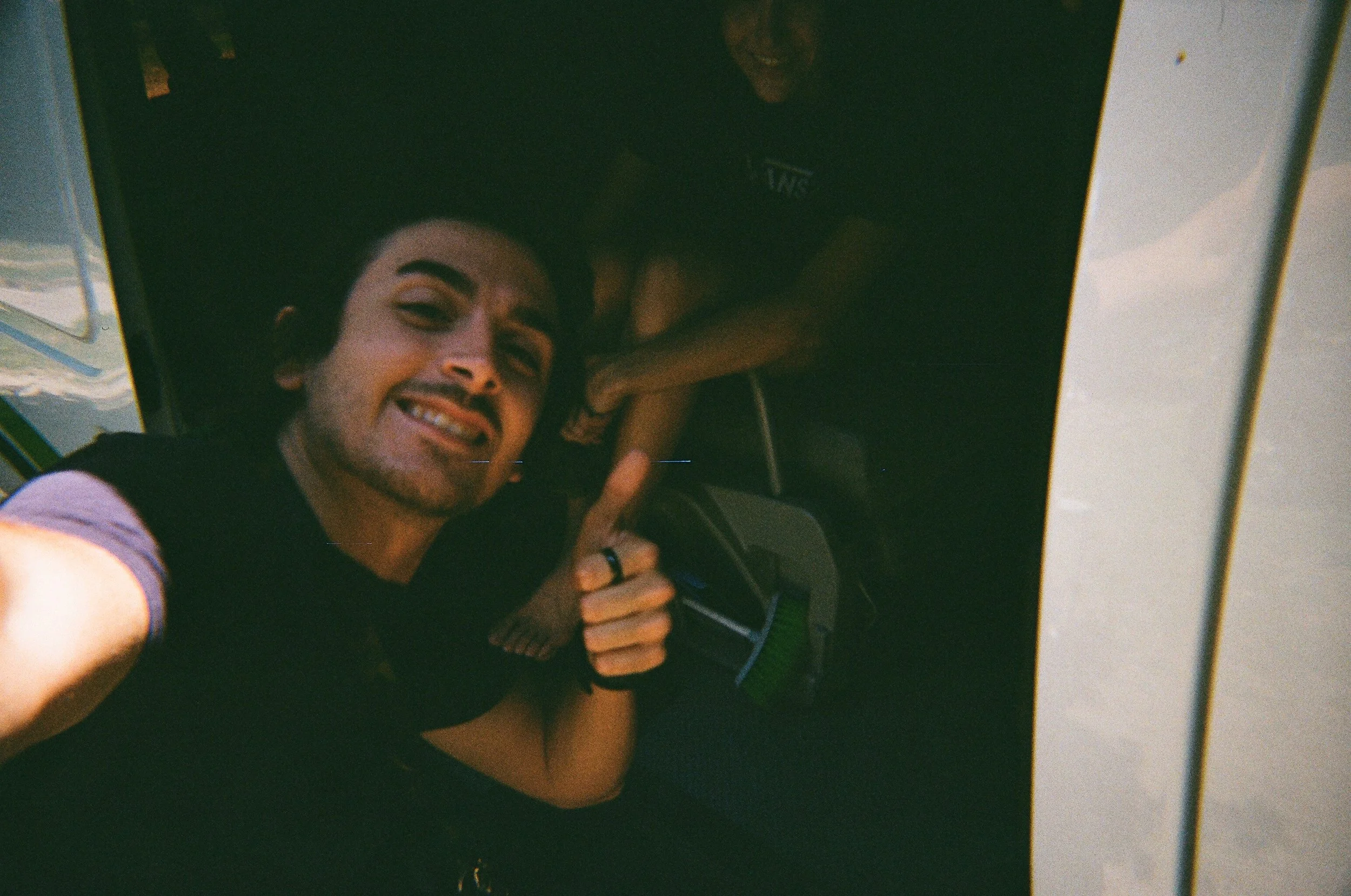 Un uomo sorridente che fa un segno di vittoria con la mano, si trova in un ambiente con un soffitto scuro e una parete bianca a destra, probabilmente in un'auto o un veicolo.