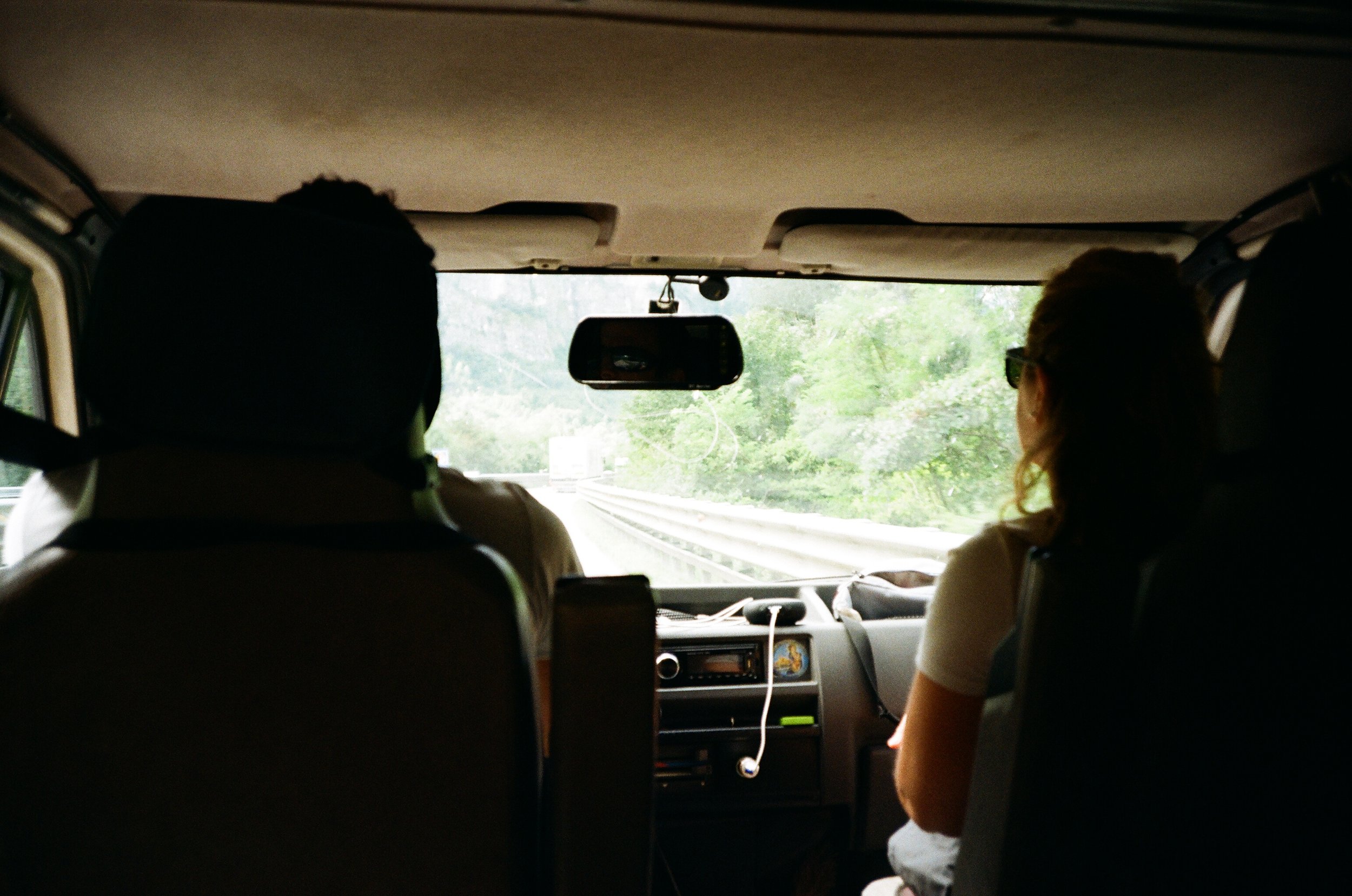 Interno di un'auto con due passeggeri, uno al volante e una donna seduta accanto, mentre viaggiano in un'area verde.