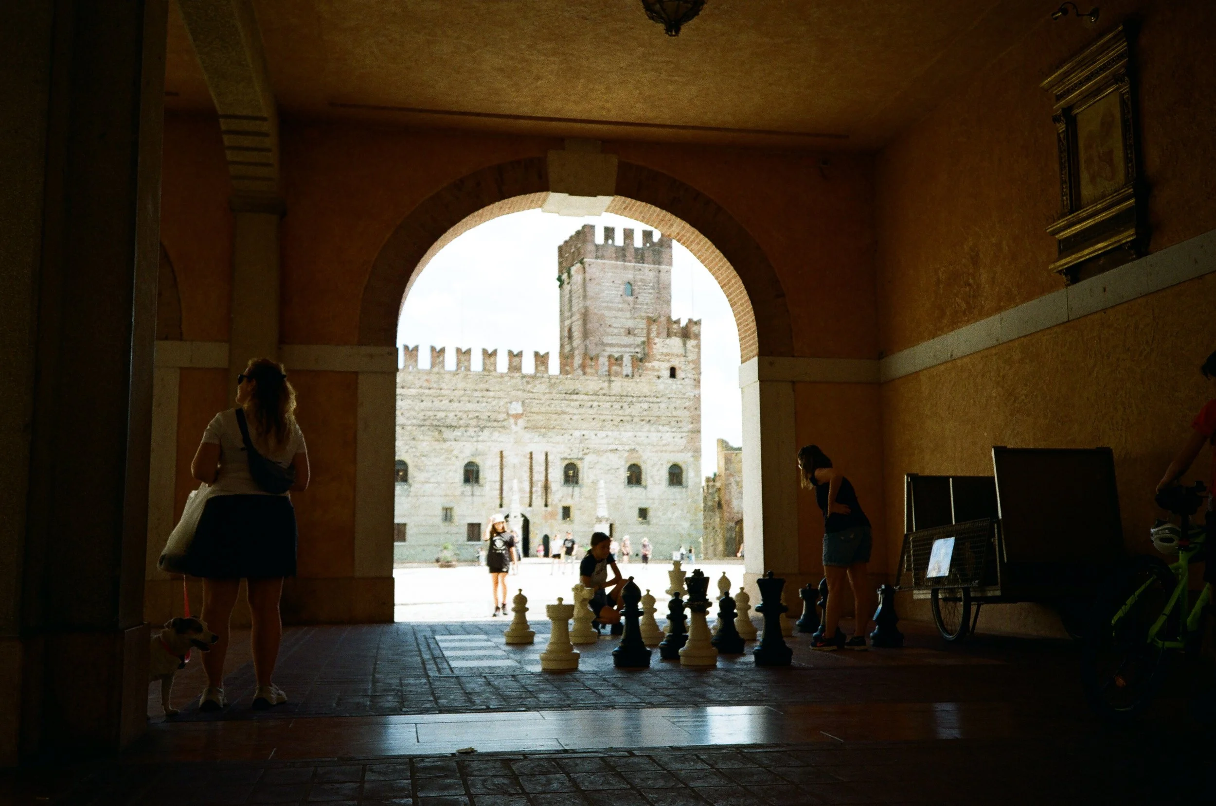 Persone all'interno di un arco osservano delle scacchiere giganti, con un castello storico visibile all'esterno.