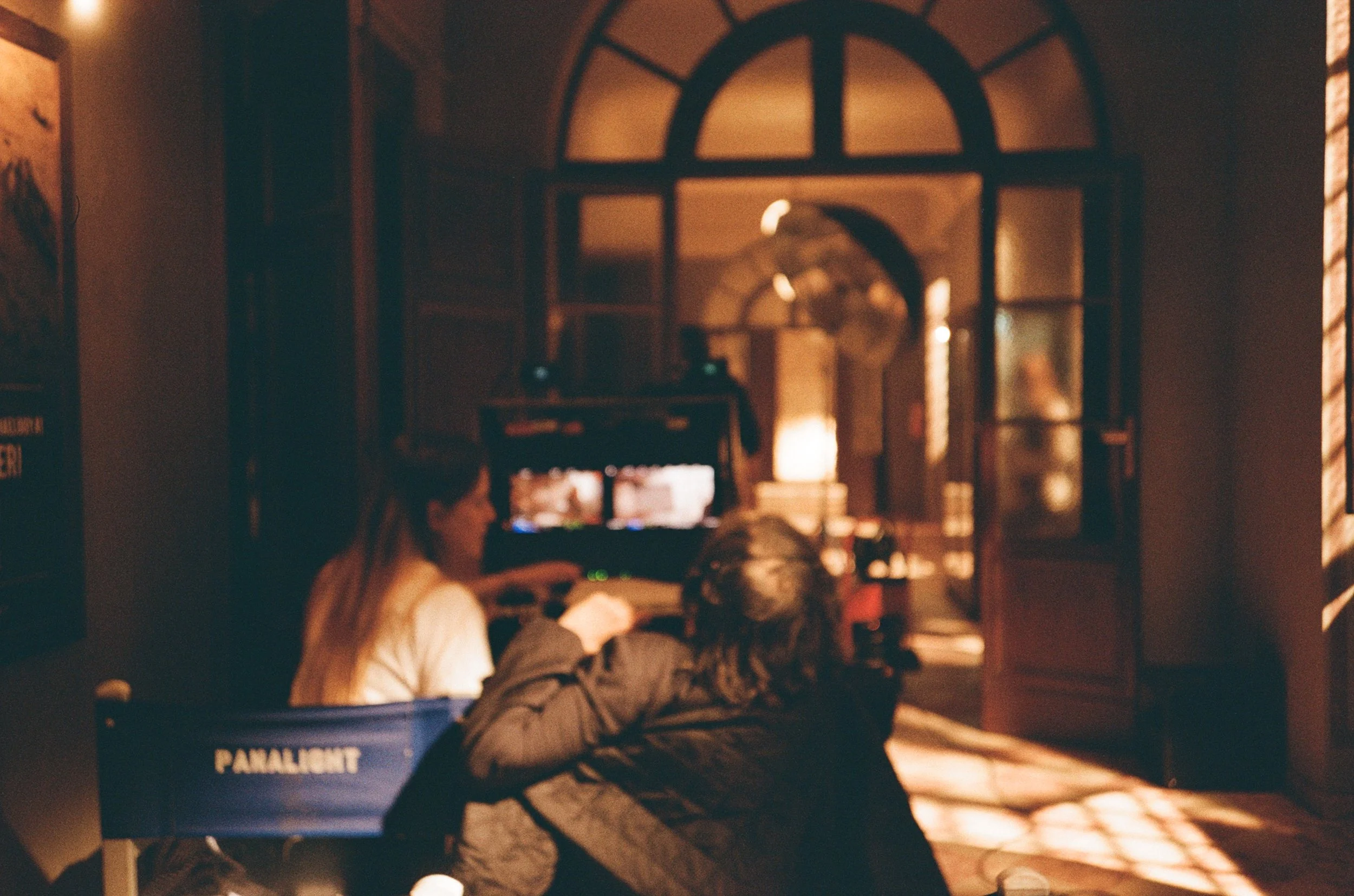 Due persone sedute in una sala scura, di fronte a un videogioco arcade, con luci calde e finestre con persiane di legno che lasciano entrare la luce.