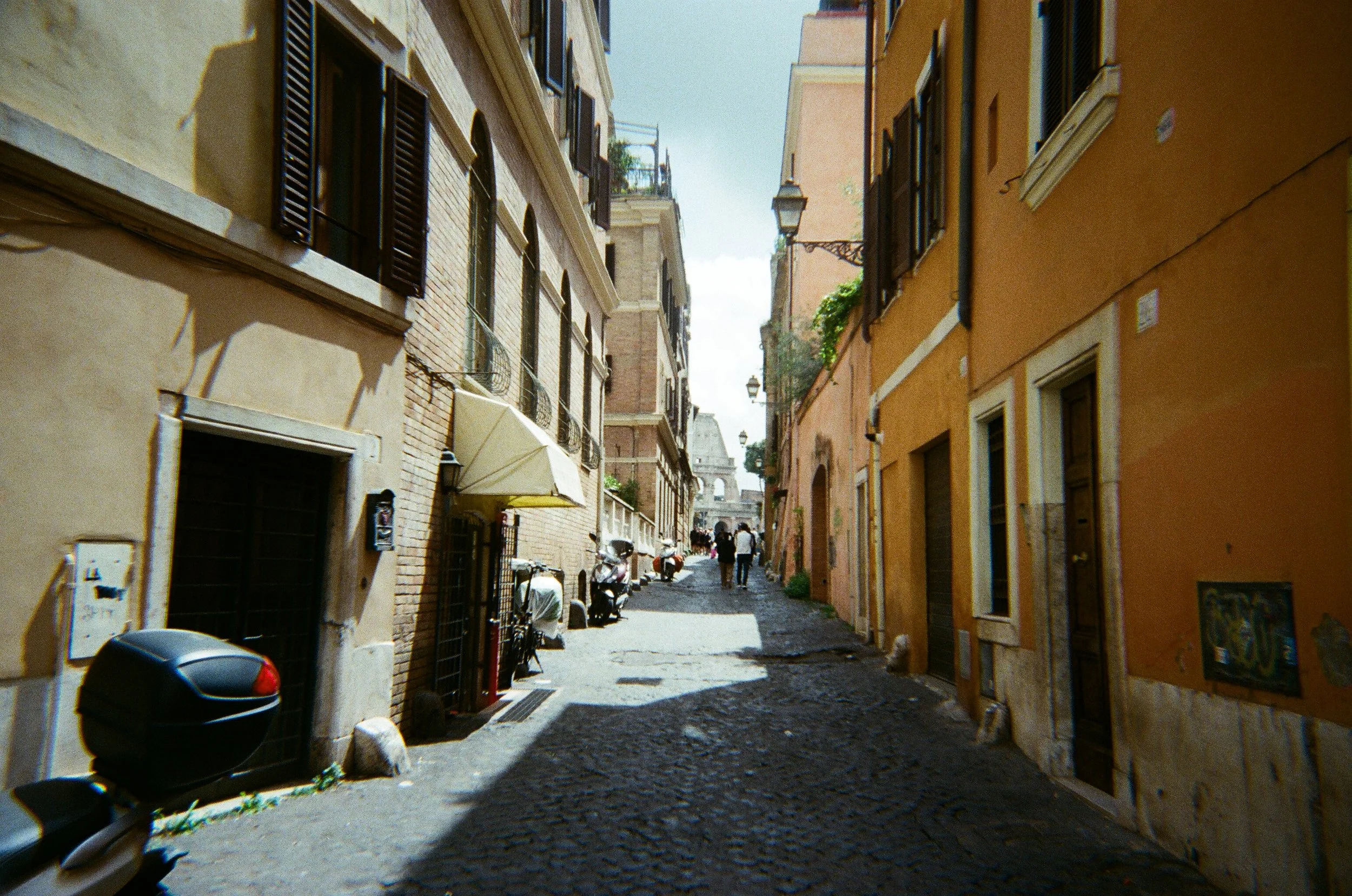 Calle stretta con edifici color ocra e beige, con finestre con persiane in legno e qualche lampione lungo la strada, con alcune persone in lontananza.