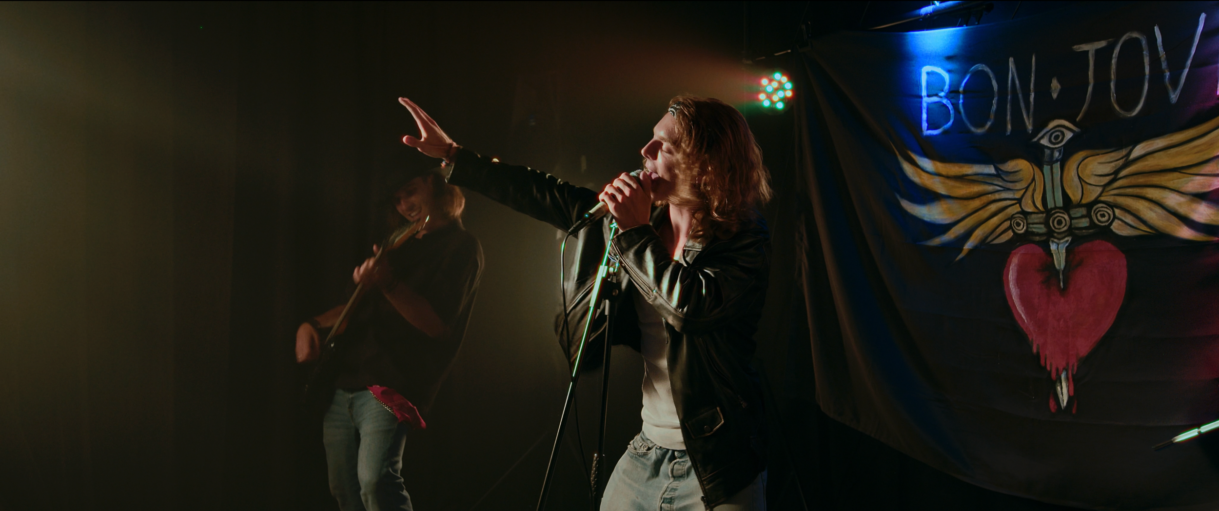 Una cantante e una chitarrista si esibiscono su un palco in un locale con luci colorate, dietro un banner con il testo 'BON' e un disegno di un cuore trafitto da un'ala con un occhio.