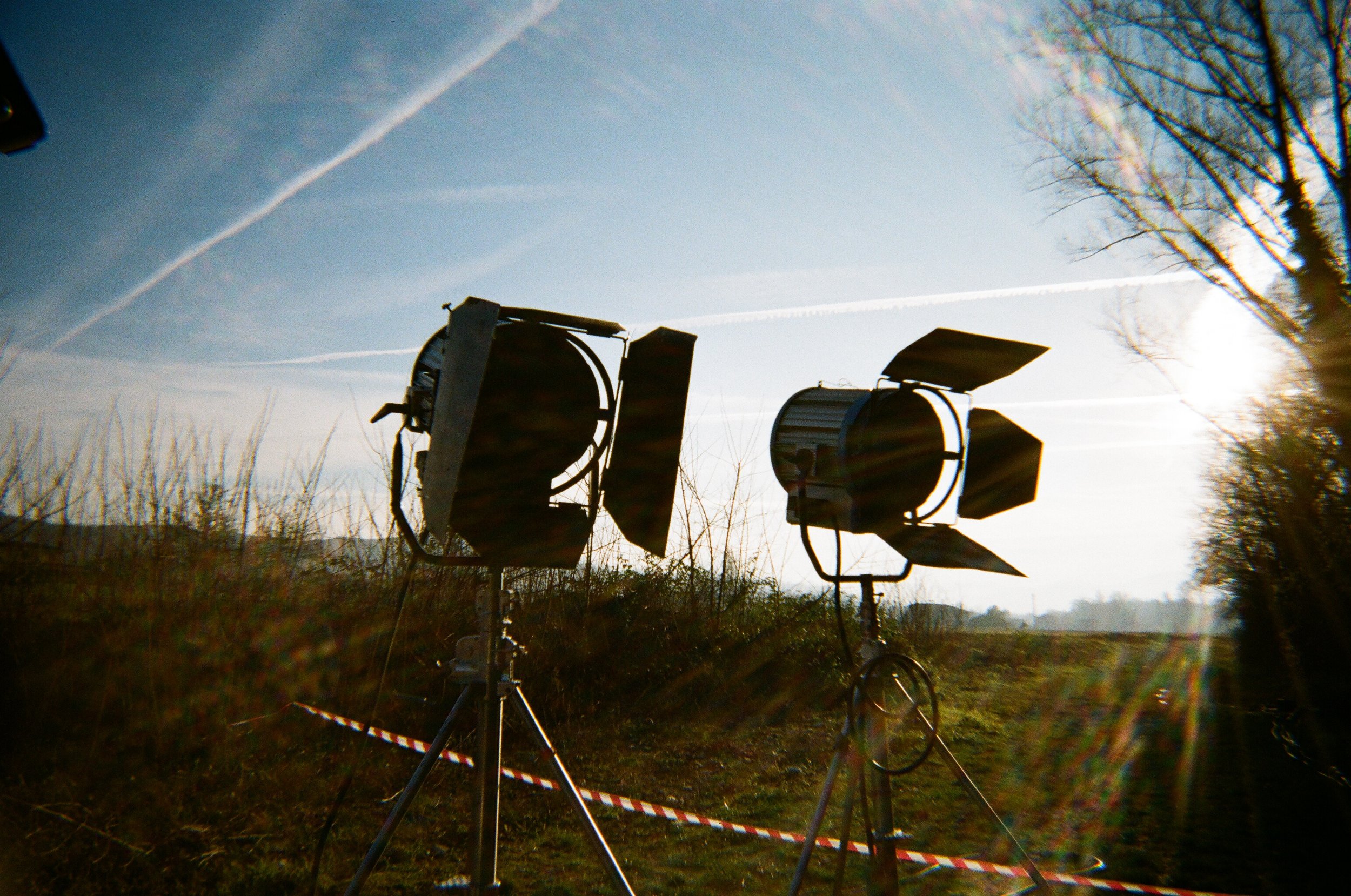 Due luci da teatro puntate verso l'alto su un terreno all'aperto con il cielo sereno e alcune nuvole e vapori di aerei in background