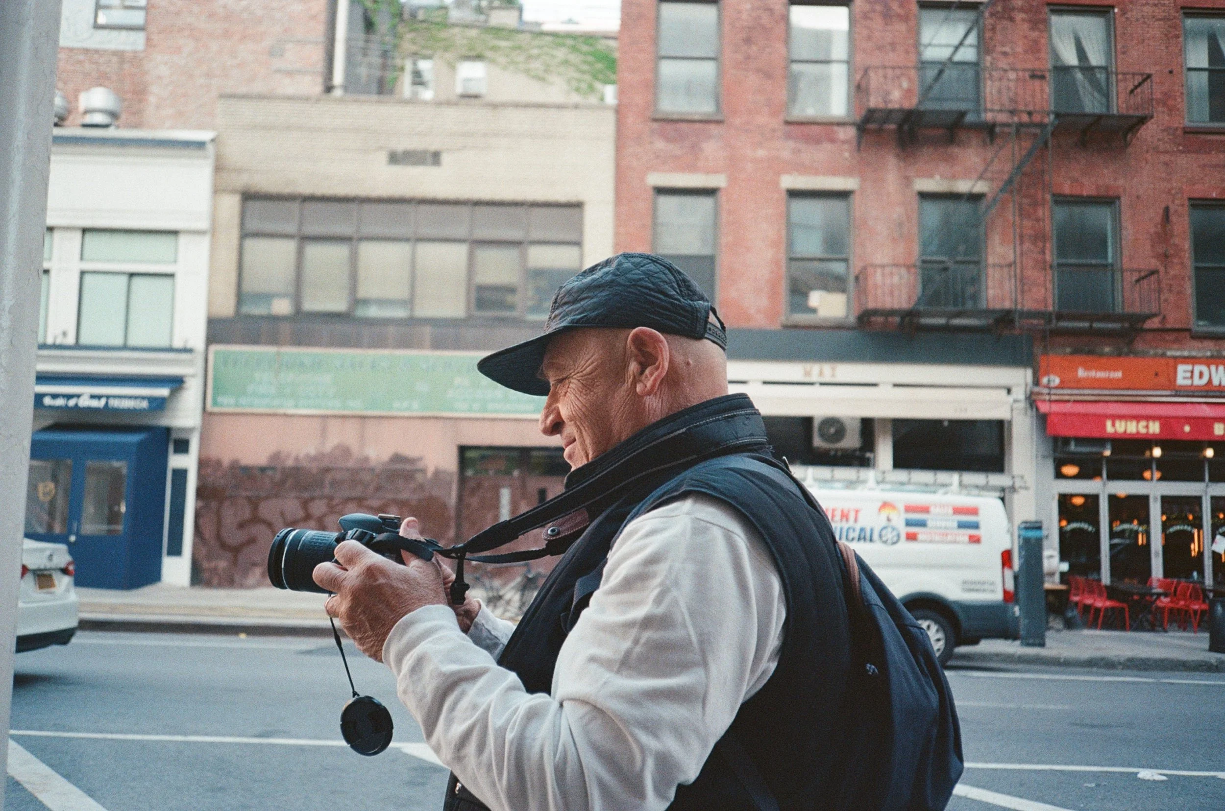 Un uomo anziano con cappellino e giacca porta uno zaino, mentre guarda e ha in mano una fotocamera in una strada urbana con edifici e veicoli sullo sfondo.