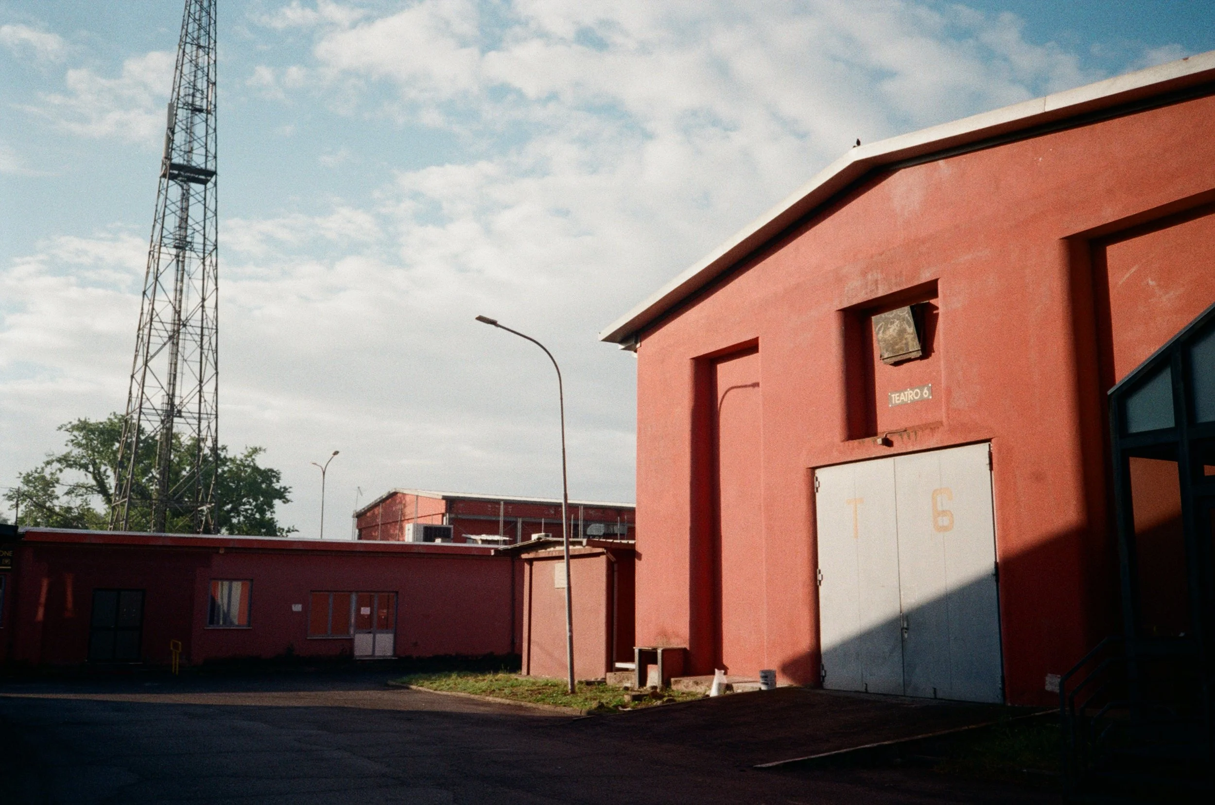 Un edificio rosso con una grande porta grigia, una piccola finestra e un cartello con 'TEATRO 6'. Sullo sfondo, un'altra struttura e un'alta torre metallica. Cielo parzialmente nuvoloso, lampioni lungo la strada.