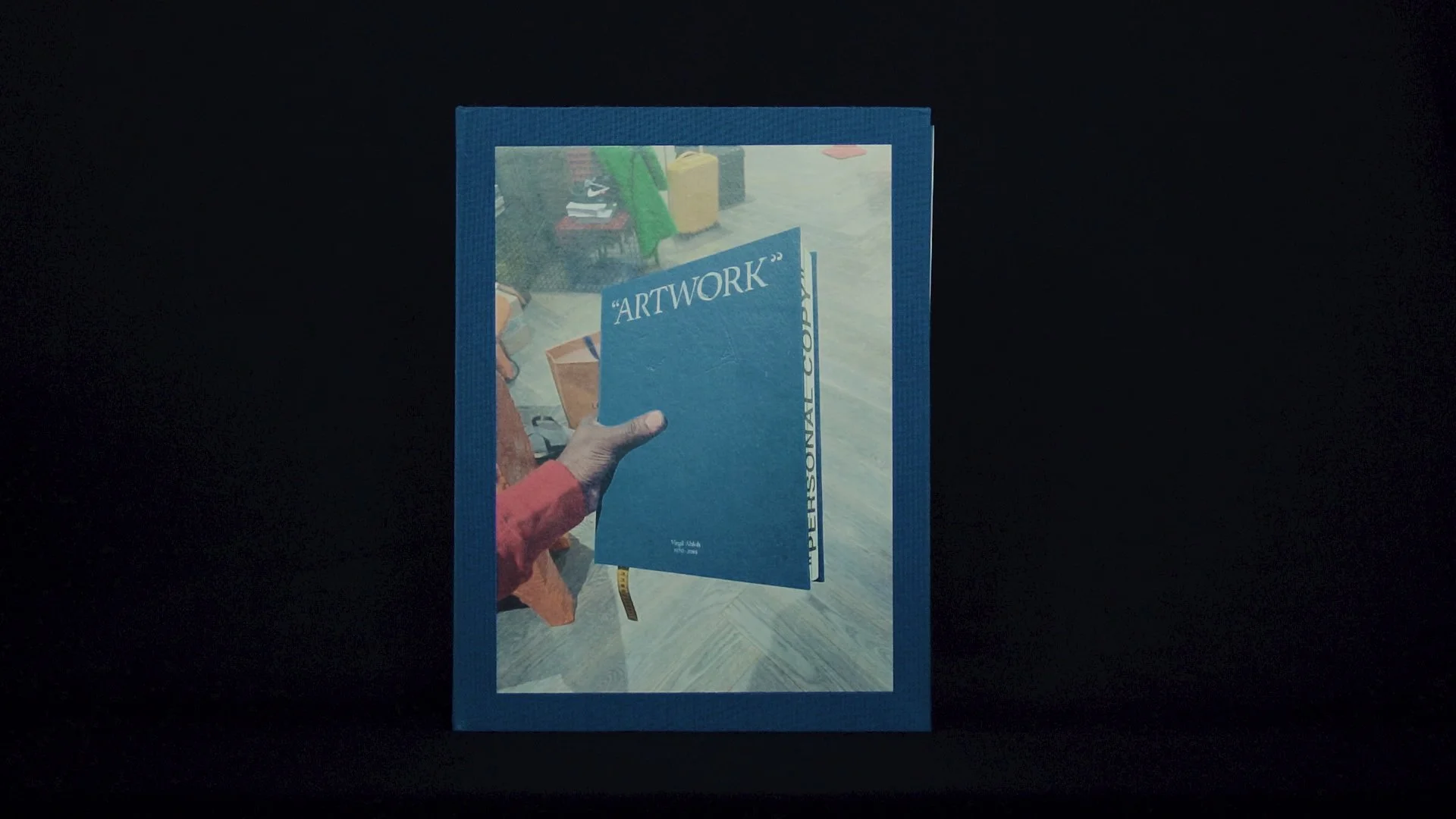 Una mano tiene una cartella blu con la parola 'ARTWORK' sopra. Sullo sfondo si vedono altre persone.