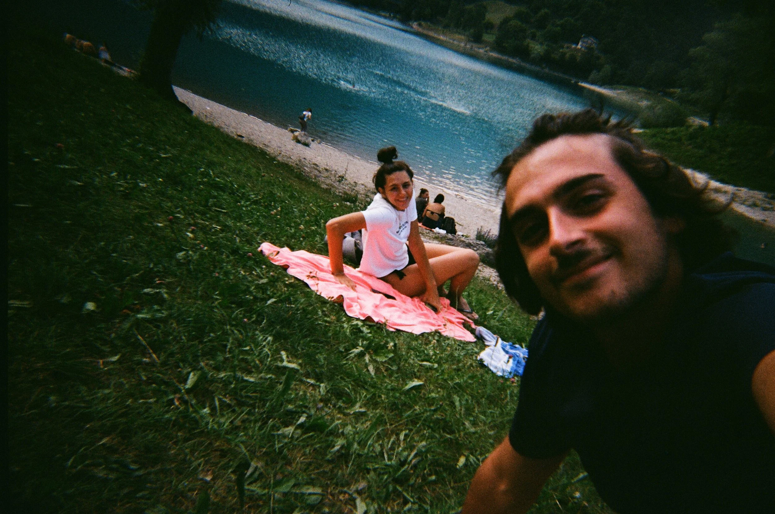 Due persone fanno un selfie vicino a un lago, con una ragazza seduta su un telo rosa e un ragazzo che si avvicina per il selfie. Sullo sfondo ci sono alcune persone che si godono la vista sulla riva del lago e alcune barche in acqua.