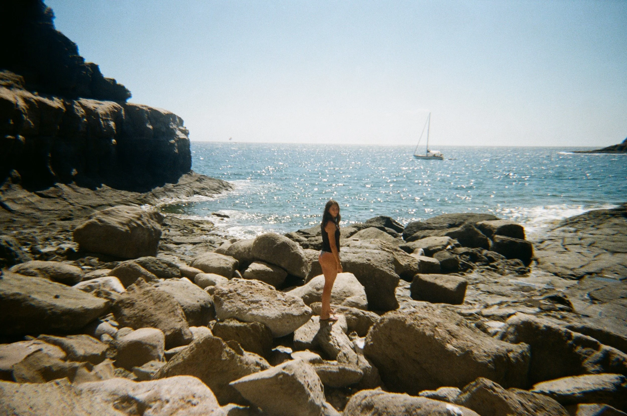 Donna in costume sulla spiaggia tra rocce con il mare e una barca a vela sullo sfondo al sole