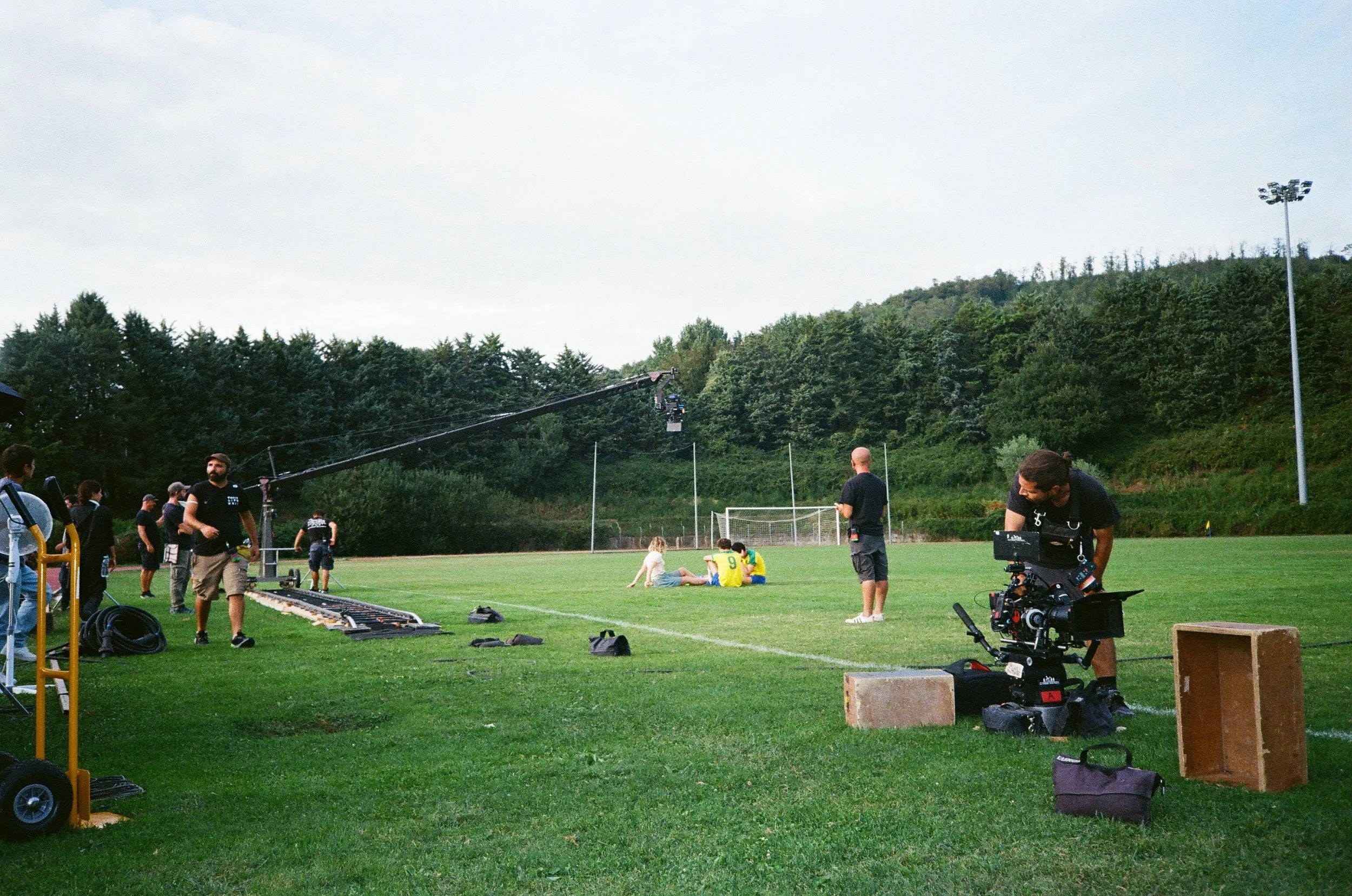 Set cinematografico sul campo sportivo con operatori e attrezzature, ripresa di scene con due attori e riflettori acceso.