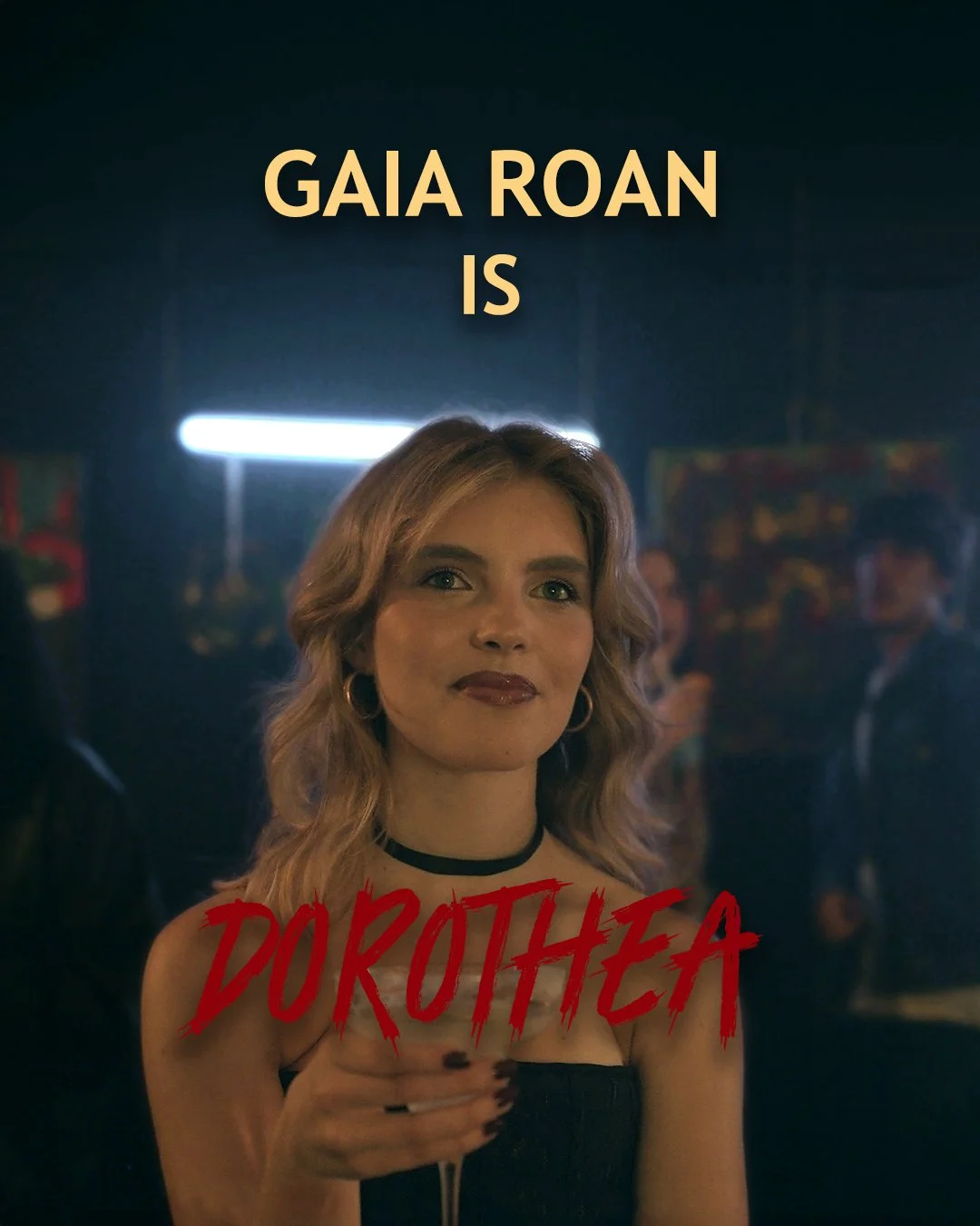 Una donna con capelli biondi ricci, trucco scuro, indossa un abito nero e una collana nera, tiene un bicchiere e sorride lievemente in un ambiente con luci soffuse. Sopra di lei c'è scritto 'GAIA ROAN IS' e in basso il titolo 'DOROTHEA' in caratteri 