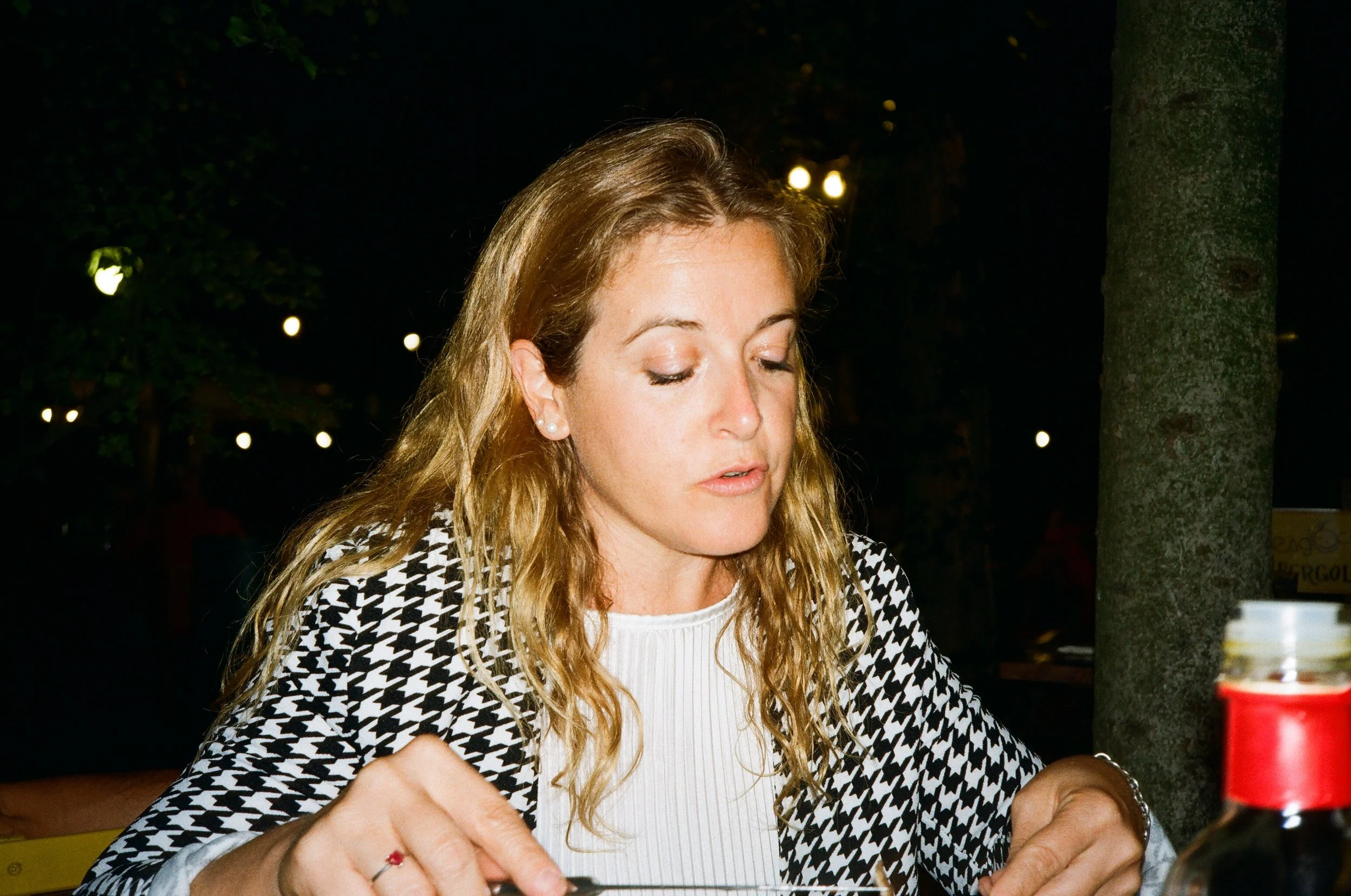 Una donna con capelli castani riccioli, vestita con una maglietta bianca e una giacca a quadri, siede all'aperto di sera con luci soffuse sullo sfondo.