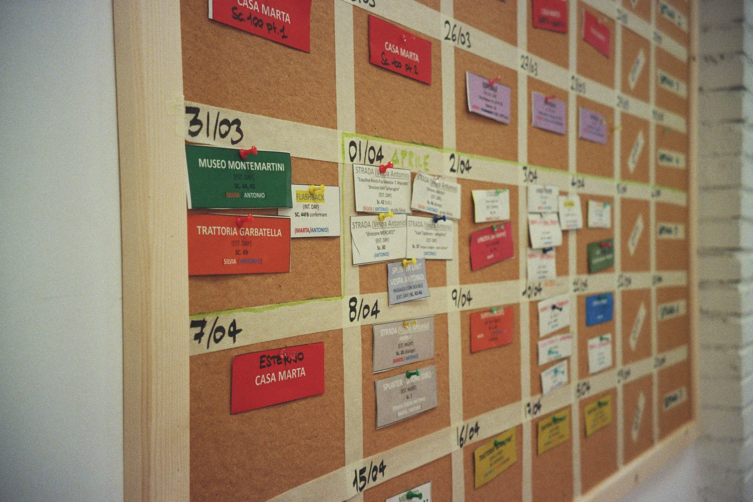 Tabellone di avvisi con note adesive colorate e biglietti attaccati, organizzato per date con riferimenti a luoghi come Museo Montemartini e Trattoria Garbatella, in un ambiente interno.