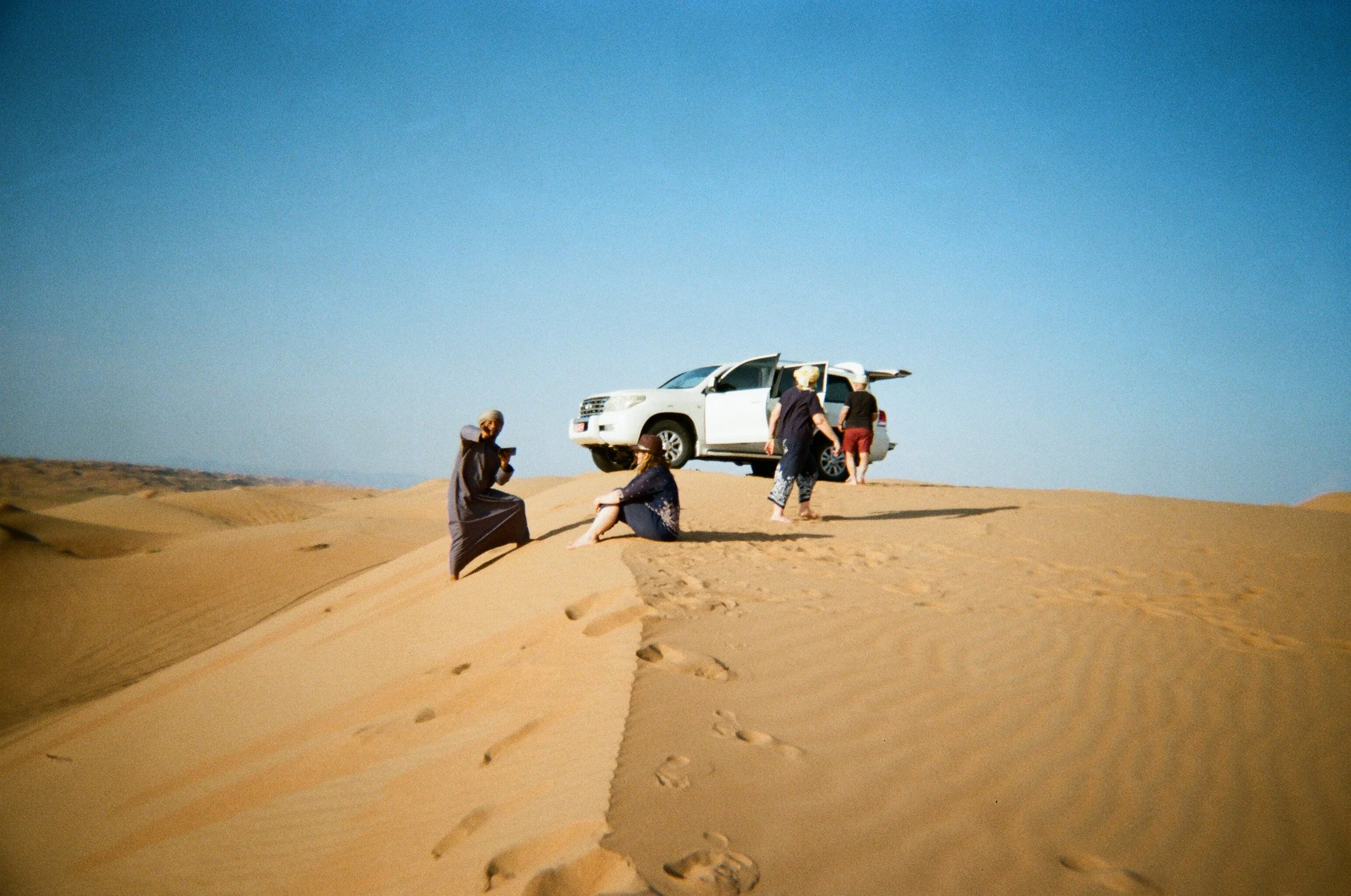 Gruppo di persone in un deserto con un'auto bianca parcheggiata su una duna di sabbia, alcune persone stanno sedute sulla sabbia e altre si avvicinano all'auto sotto un cielo azzurro.