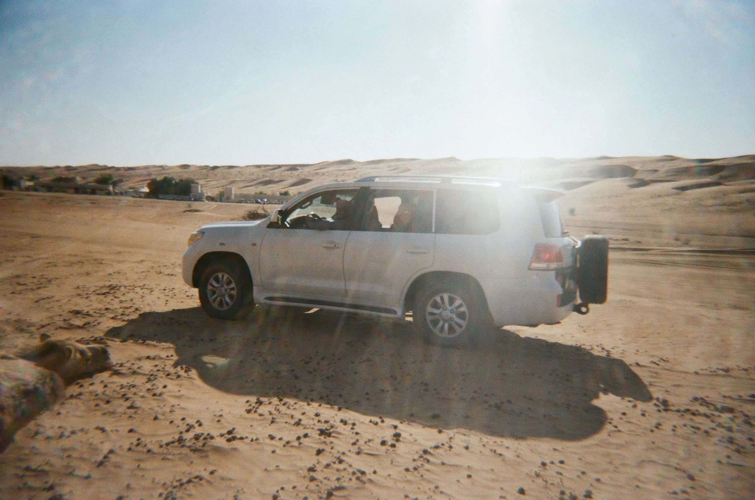 Auto in un deserto sabbioso con dune al fondo, motivo per cui l'auto è parzialmente alloggiata nel terreno sabbioso, con due persone all'interno.