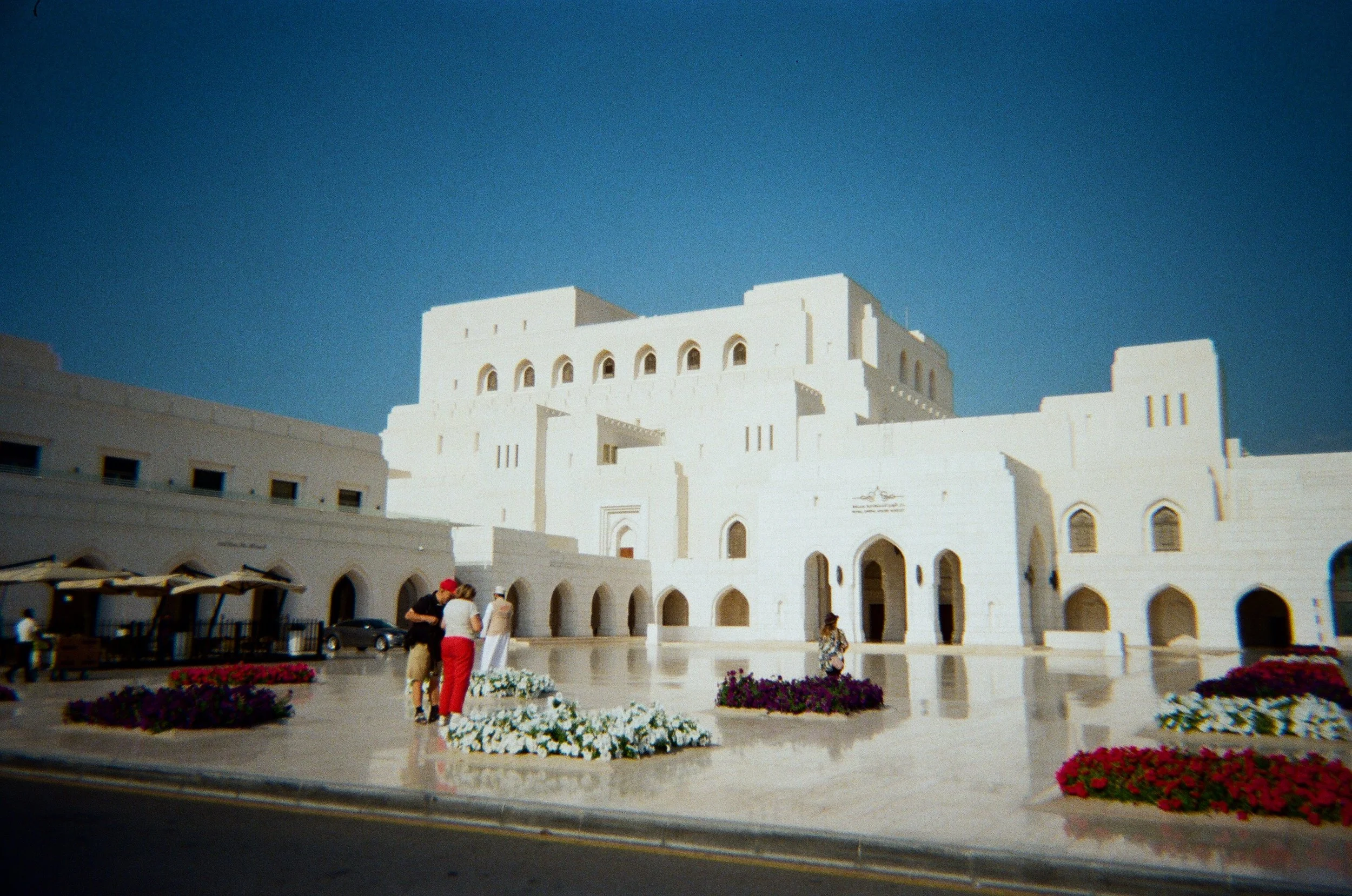 Edificio bianco in stile architettonico tradizionale con archi e varie finestre, campi di fiori colorati e persone che si aggirano all'esterno, sotto un cielo blu.