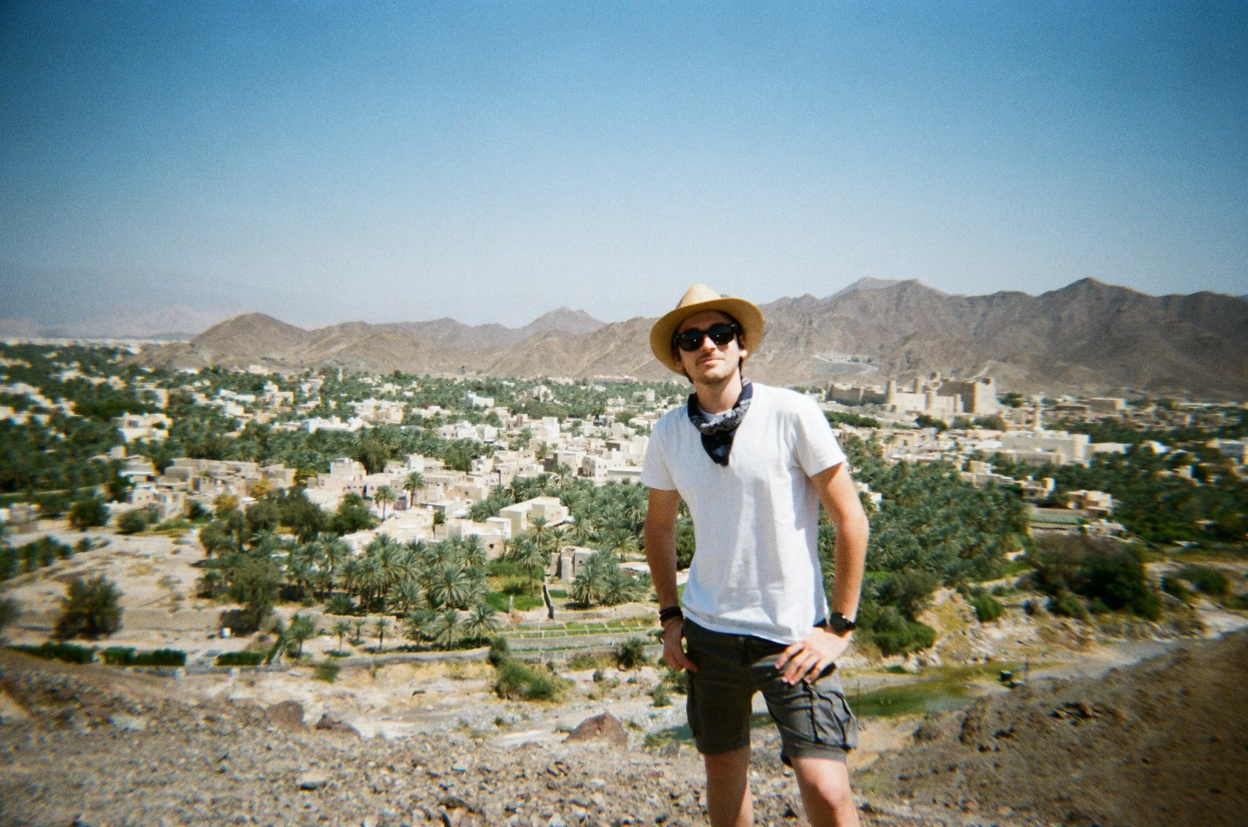 Un uomo con cappello di paglia, occhiali scuri, maglietta bianca, pantaloncini e braccialetti, si trova davanti a una vista panoramica di un'oasi con alberi di palme e edifici in una valle desertica, con montagne sullo sfondo.