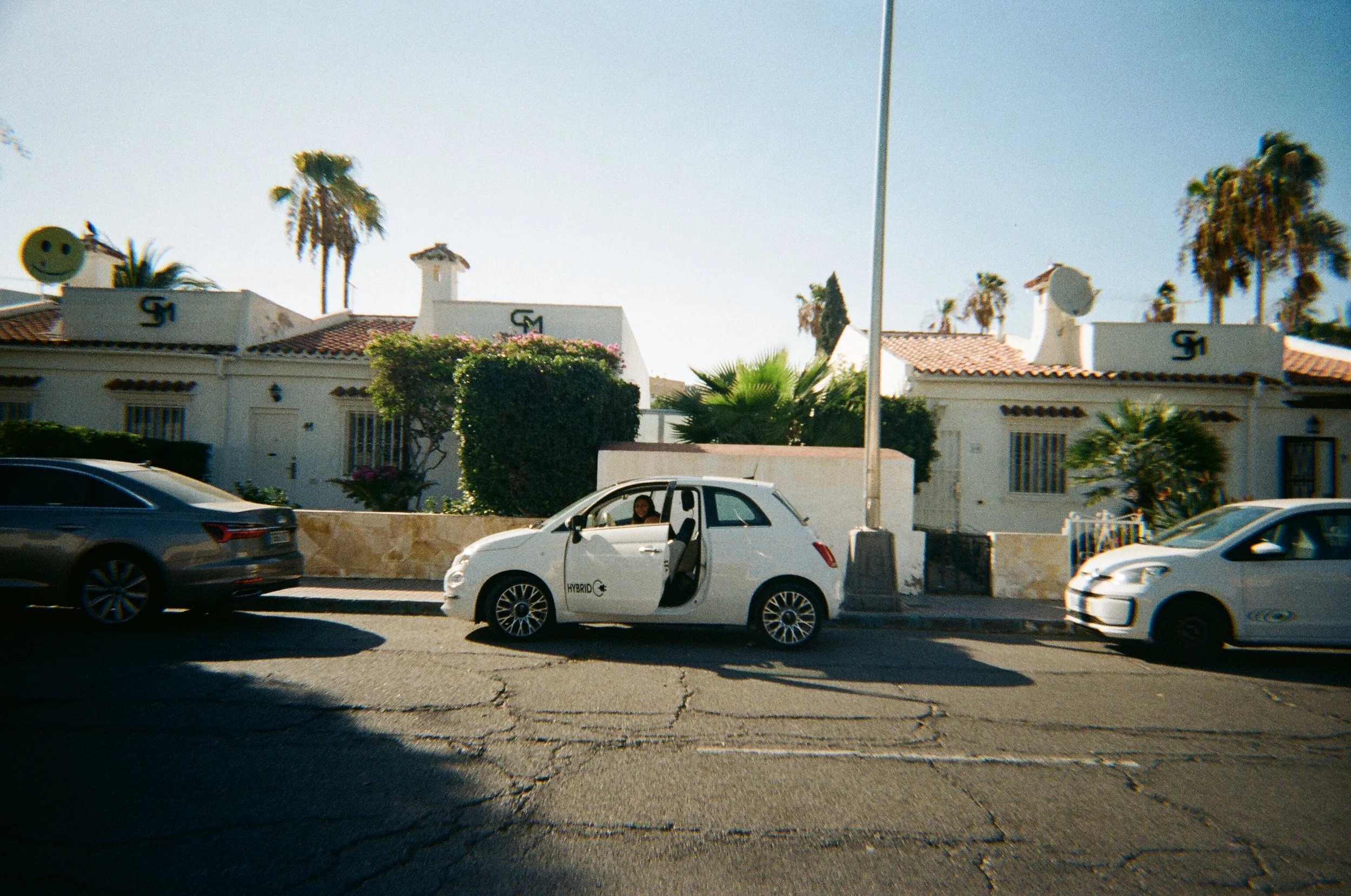 Auto ibrida bianca parcheggiata sul marciapiede, con una donna seduta al volante, di fronte a case bianche con tetti di tegole rosse e palme sullo sfondo, in una giornata di sole.