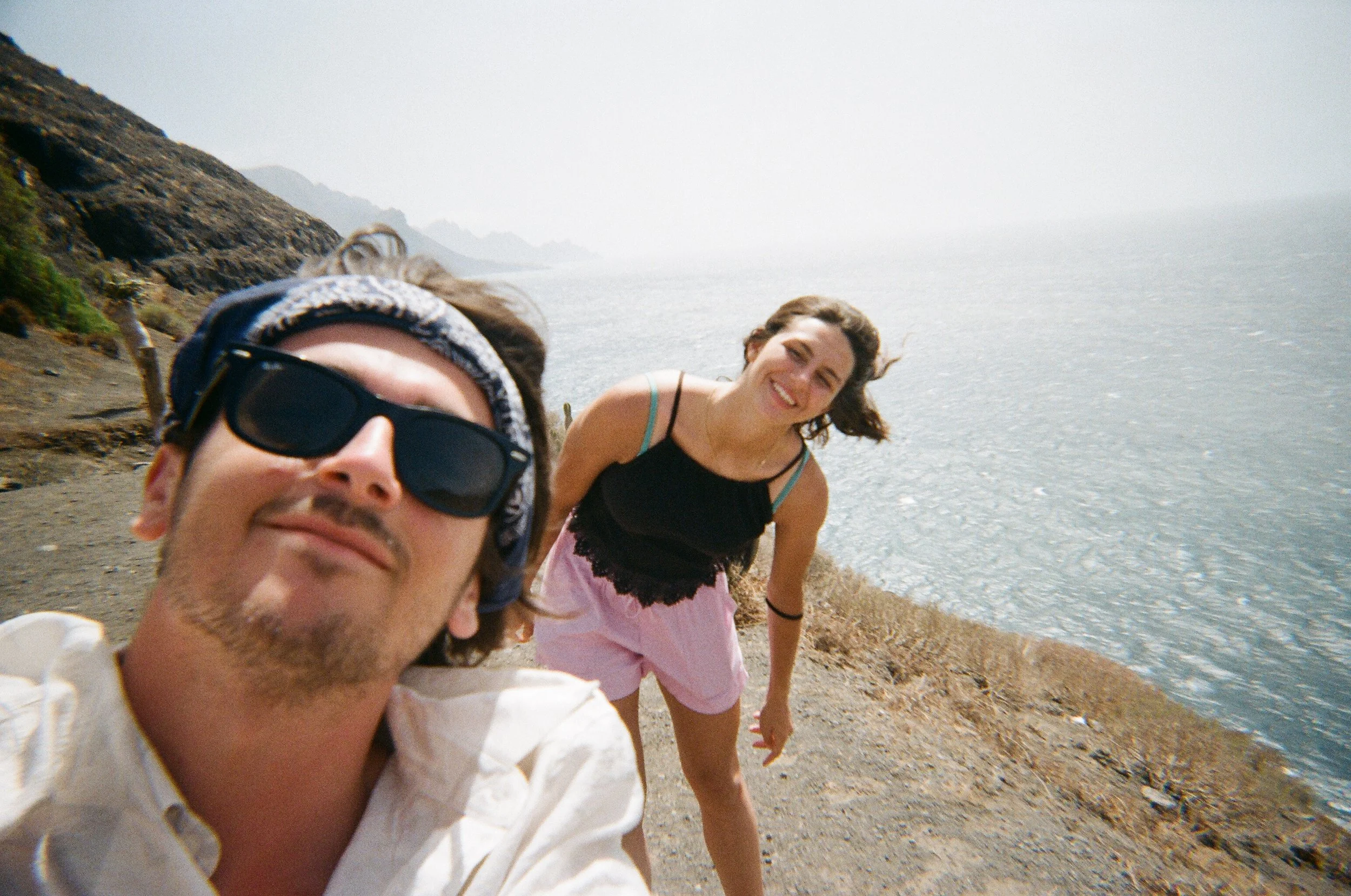 Due persone, un uomo in primo piano con occhiali da sole e bandana in testa, e una donna dietro di lui che sorride, sulla costa di un mare in una giornata ventosa.