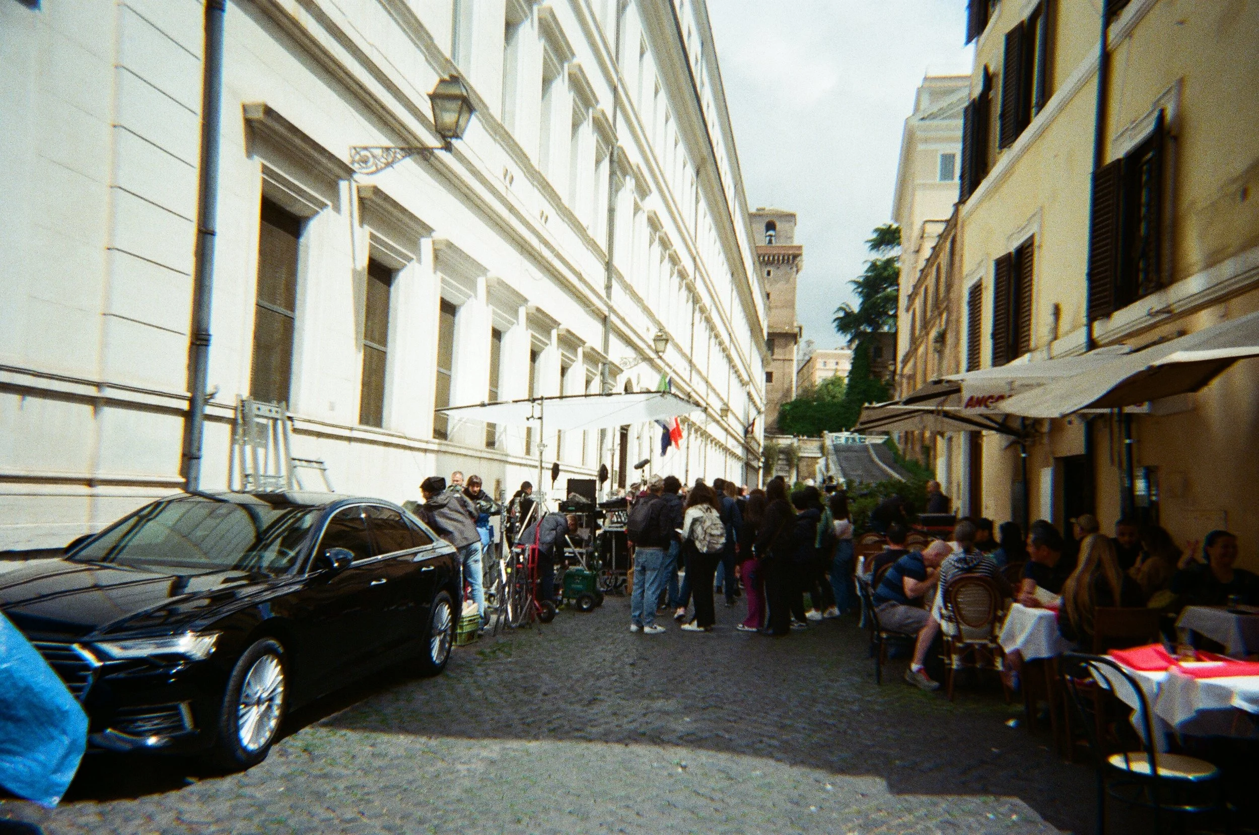 Ragazza con camicia a quadri seduta a un tavolo di un ristorante all'aperto con tavoli e sedie di fronte a un edificio di stile classico, in una strada affollata di persone.