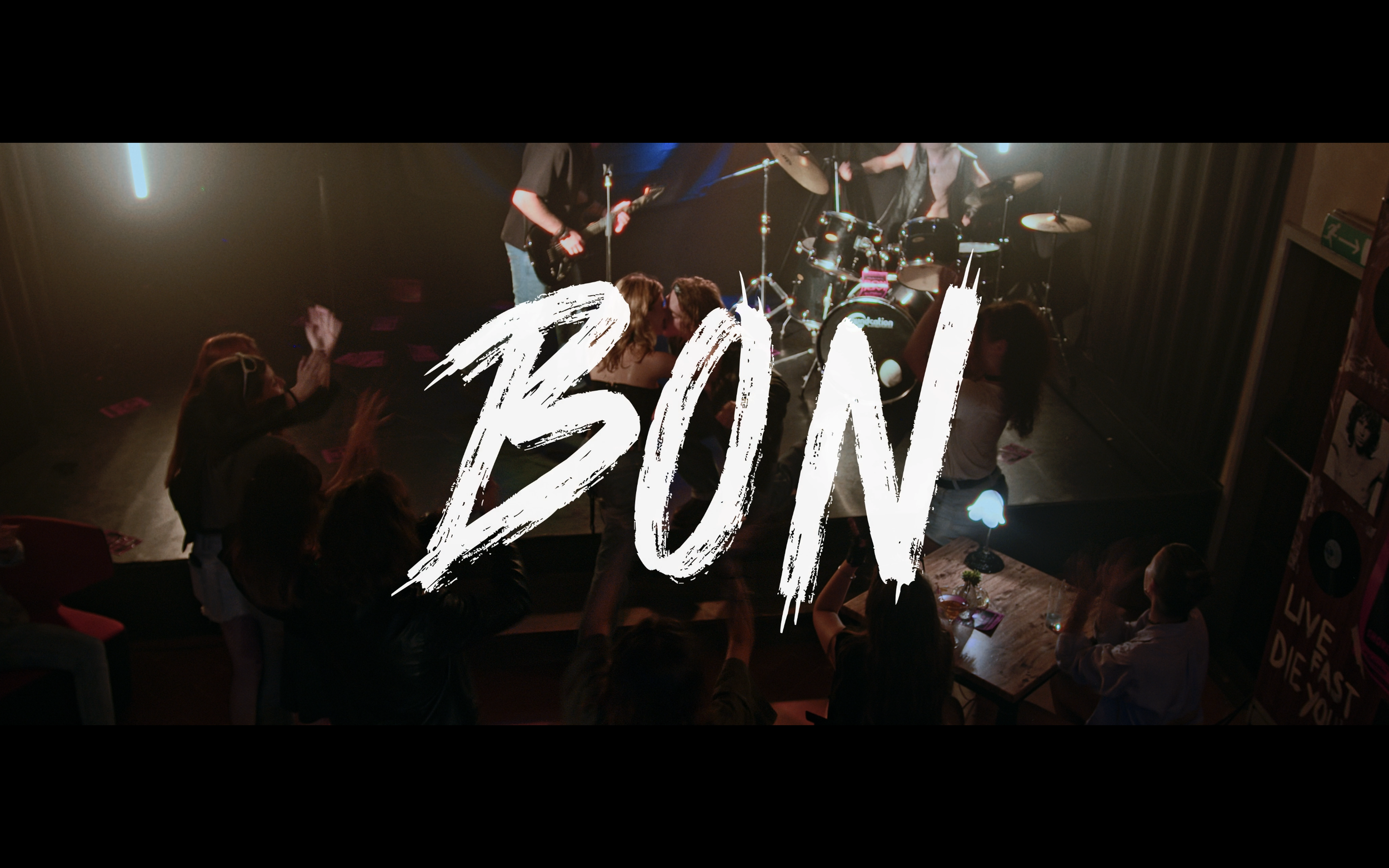Una scena di un concerto dal vivo con un pubblico che si diverte davanti a un palco con una band che suona. La parola 'BION' scritta in grande al centro dell'immagine.