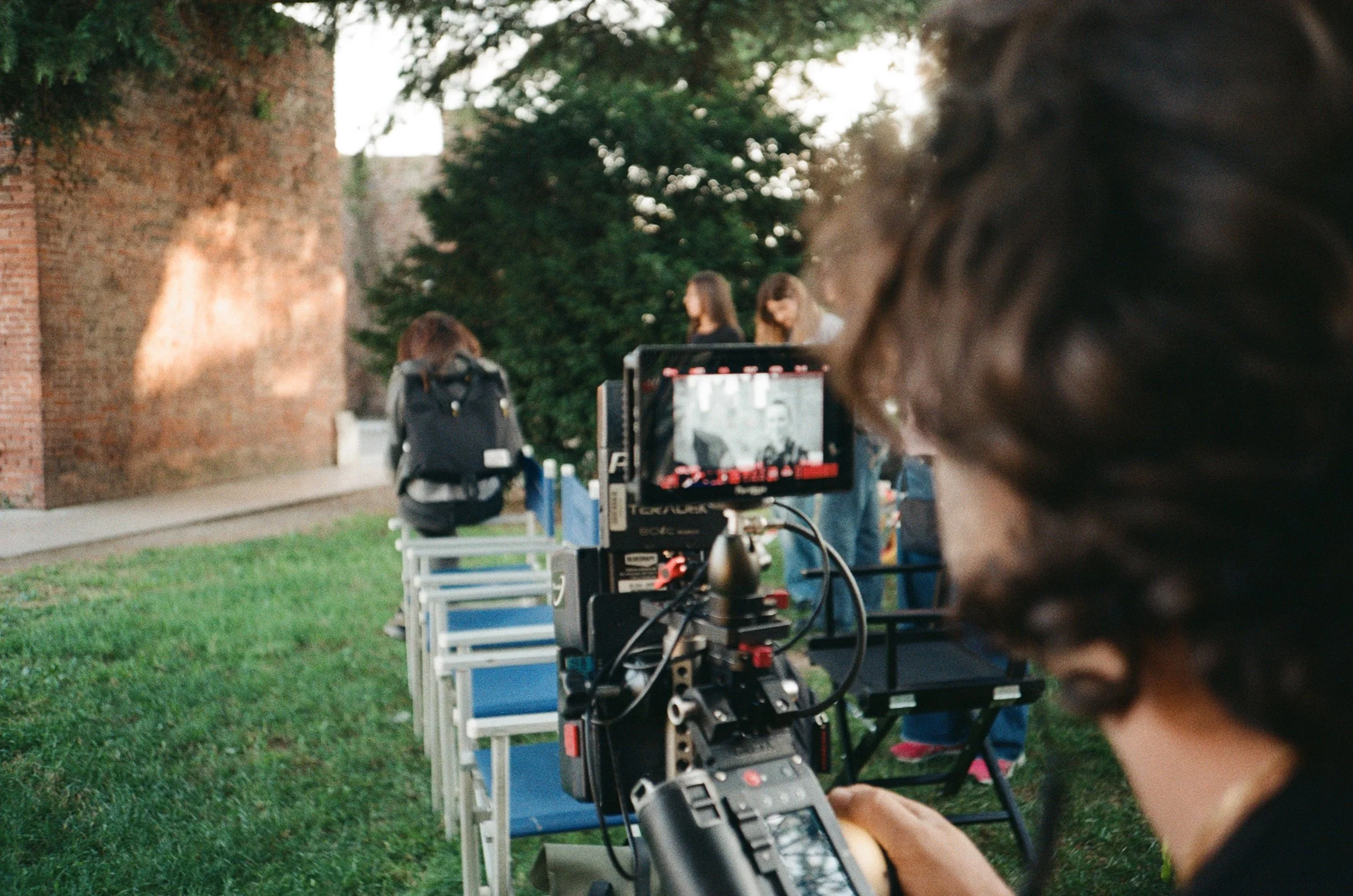 Operatore di riprese con videocamera professionale al lavoro all'aperto davanti a un gruppo di donne sedute su sedie, in un'area con albero e muro di mattoni.