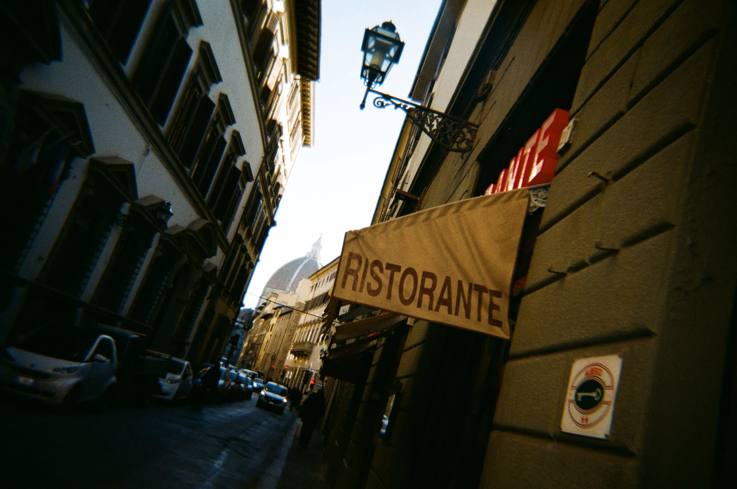 Immagine di una stretta strada cittadina con edifici storici, un cartello che dice "Ristorante" e alcune auto parcheggiate lungo la strada.