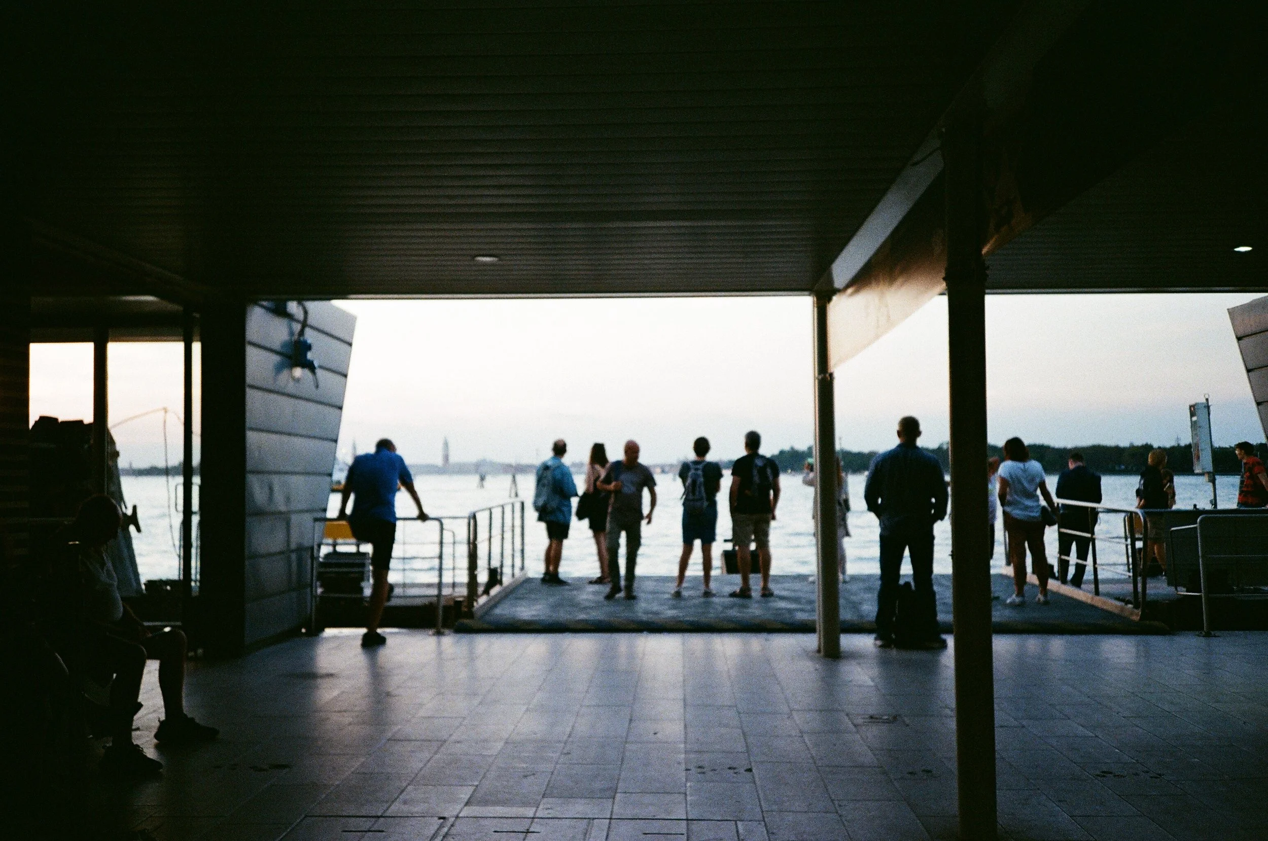 Persone che si affacciano su un molo vicino a un fiume, viste dall’interno di una struttura coperta, con le silhouette di alcune persone sedute e altre in piedi che osservano il panorama al tramonto.
