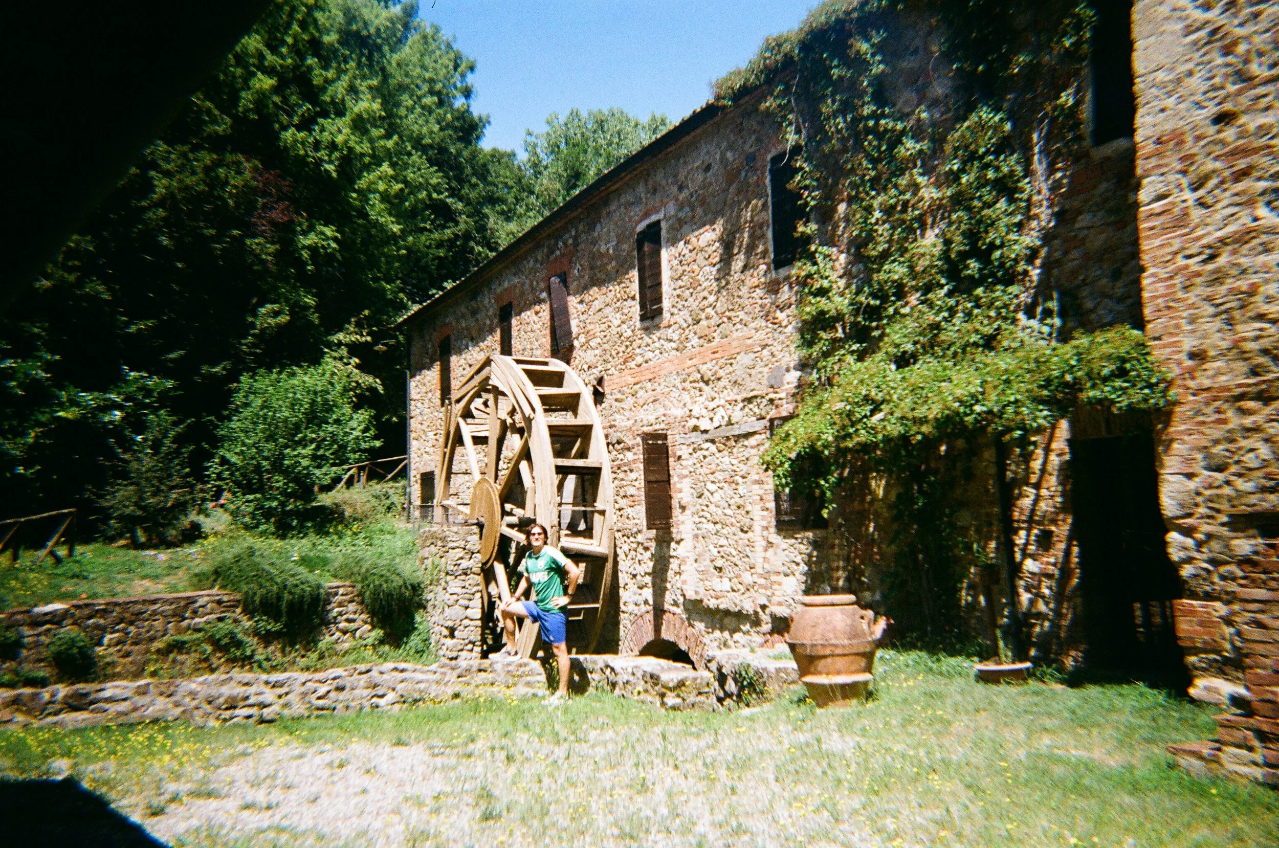 Una donna in posa davanti a un mulino ad acqua di pietra, con un edificio in pietra a due piani sullo sfondo e vegetazione rigogliosa, sotto un cielo blu soleggiato.