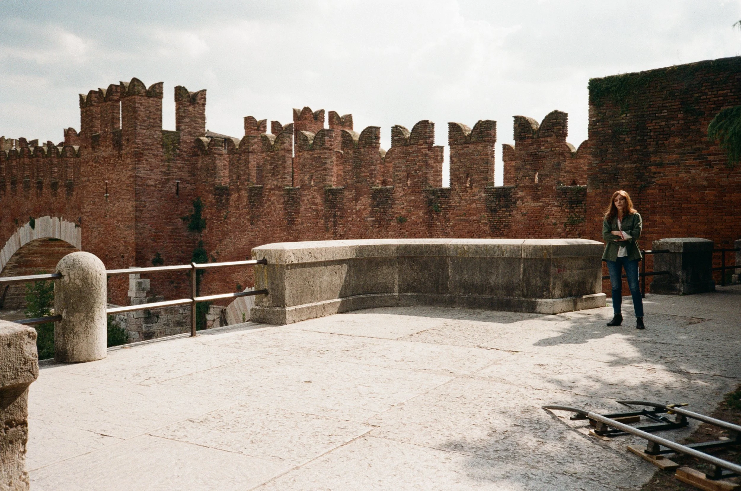 Donna con giacca verde scuro e jeans in piedi vicino a un muro di mattoni rossi con mura medievali sullo sfondo.