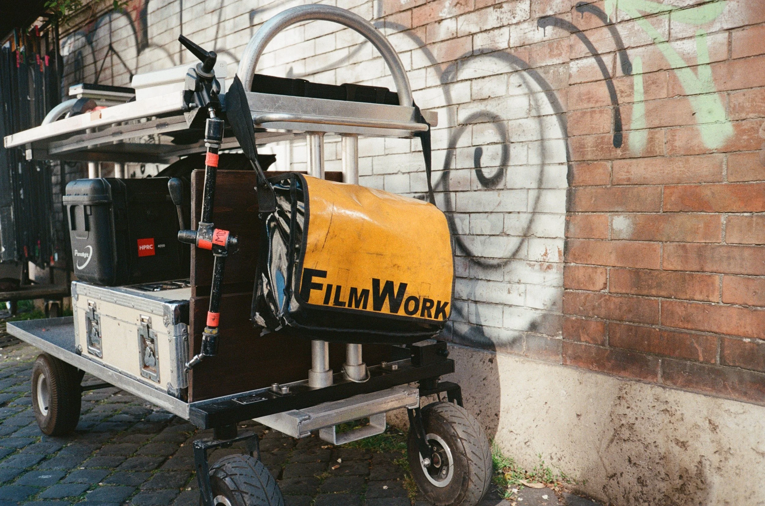 Carrello per riprese cinematografiche con attrezzature, parcheggiato su ciottolato davanti a un muro di mattoni con graffiti, con una borsa gialla con scritto 'FilmWork' appesa al lato.