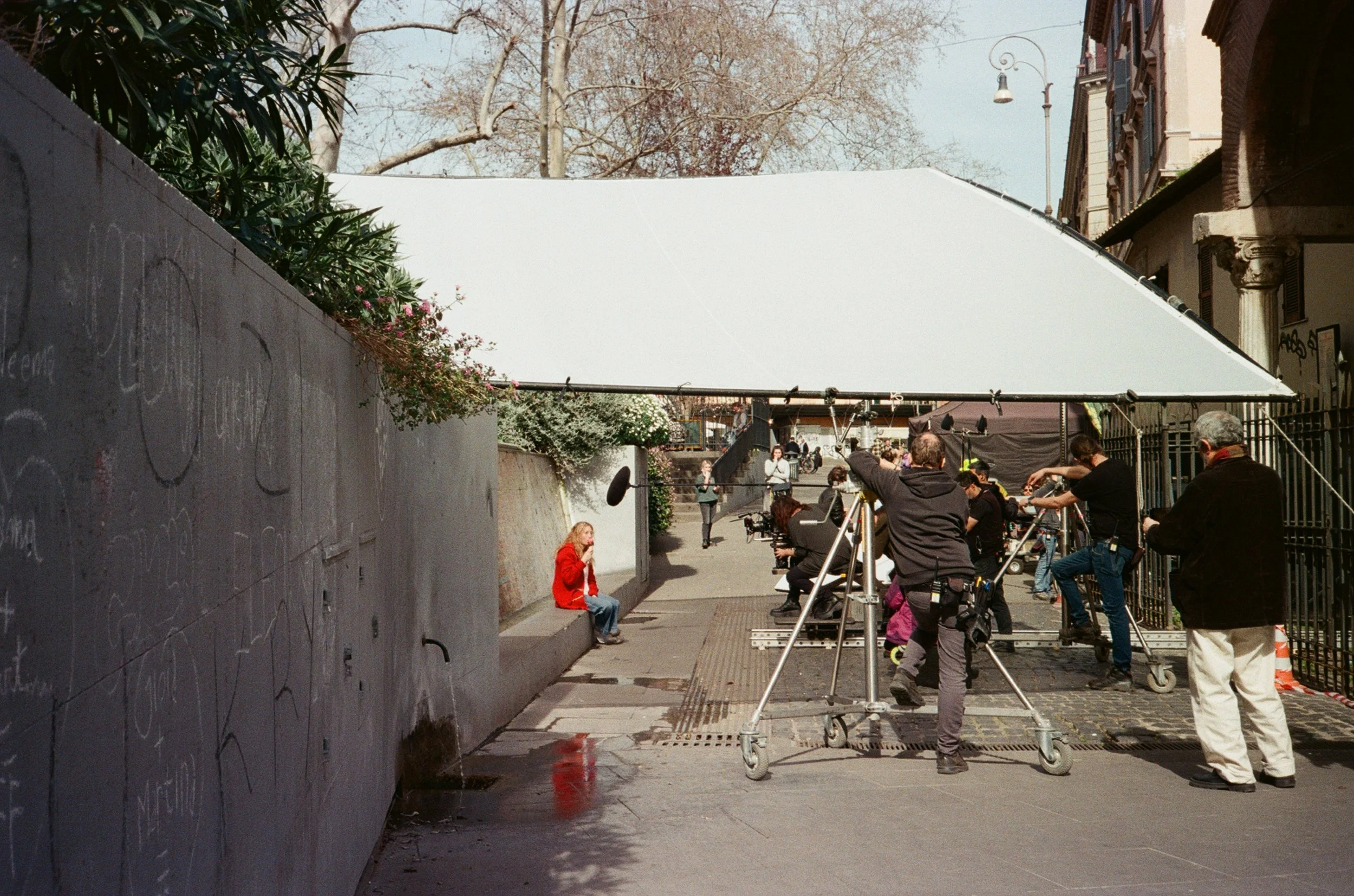 Set di riprese cinematografiche all'aperto con un team di persone intorno a una donna seduta su una panchina rossa in un vicolo cittadino, con telecamere e luci professionali, sotto un grande ombrellone bianco.