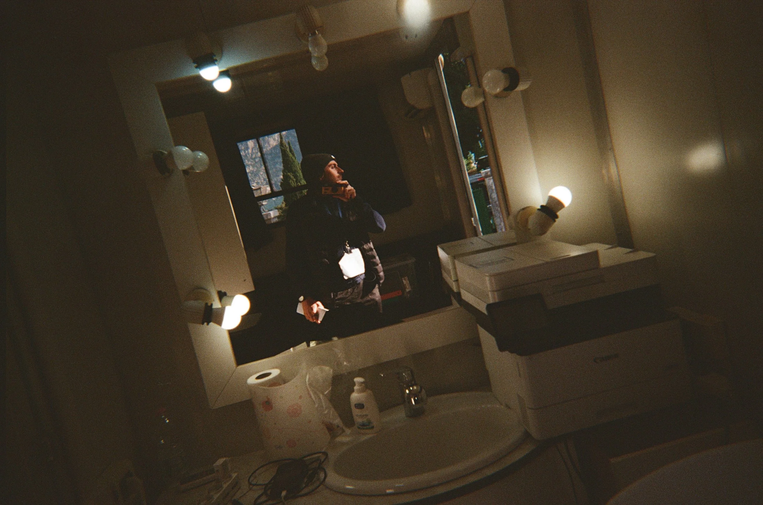 Specchio da trucco con luci, lavabo con sapone e carta igienica, mensola con scatole, in una stanza illuminata da luci calde.