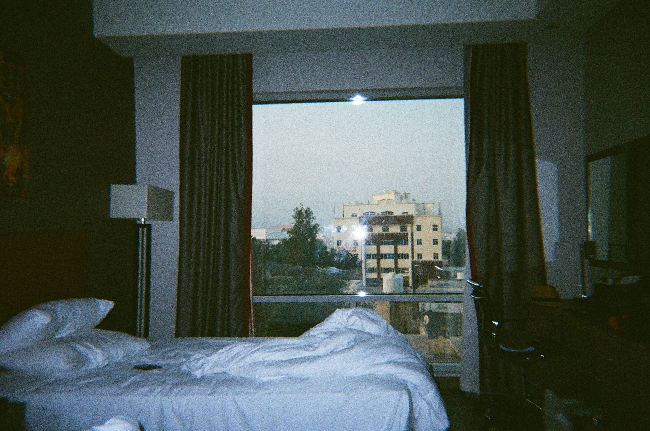 Camera cabina con letto non rifinito, tende colorate, lampada su un lato e vista su edifici e strada nell'era del tramonto.