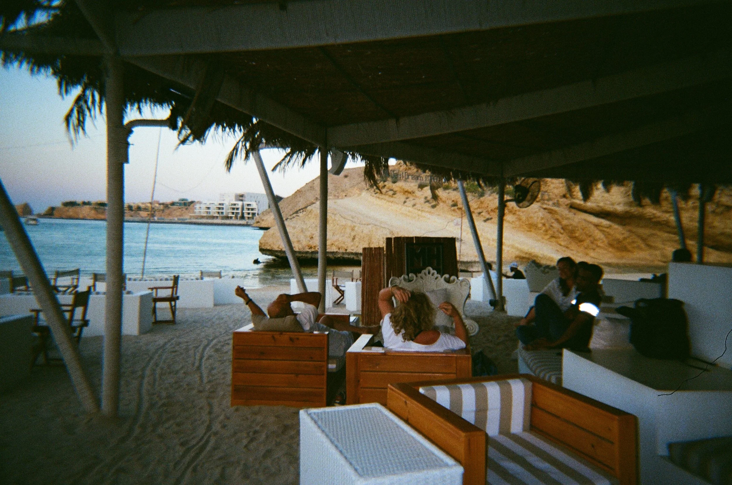 Sotto una tettoia in stile spiaggia, persone sedute su mobili in legno e stoffa, spettando il tramonto sulla spiaggia vicino a un mare calmo, con scogli e edifici in fondo.
