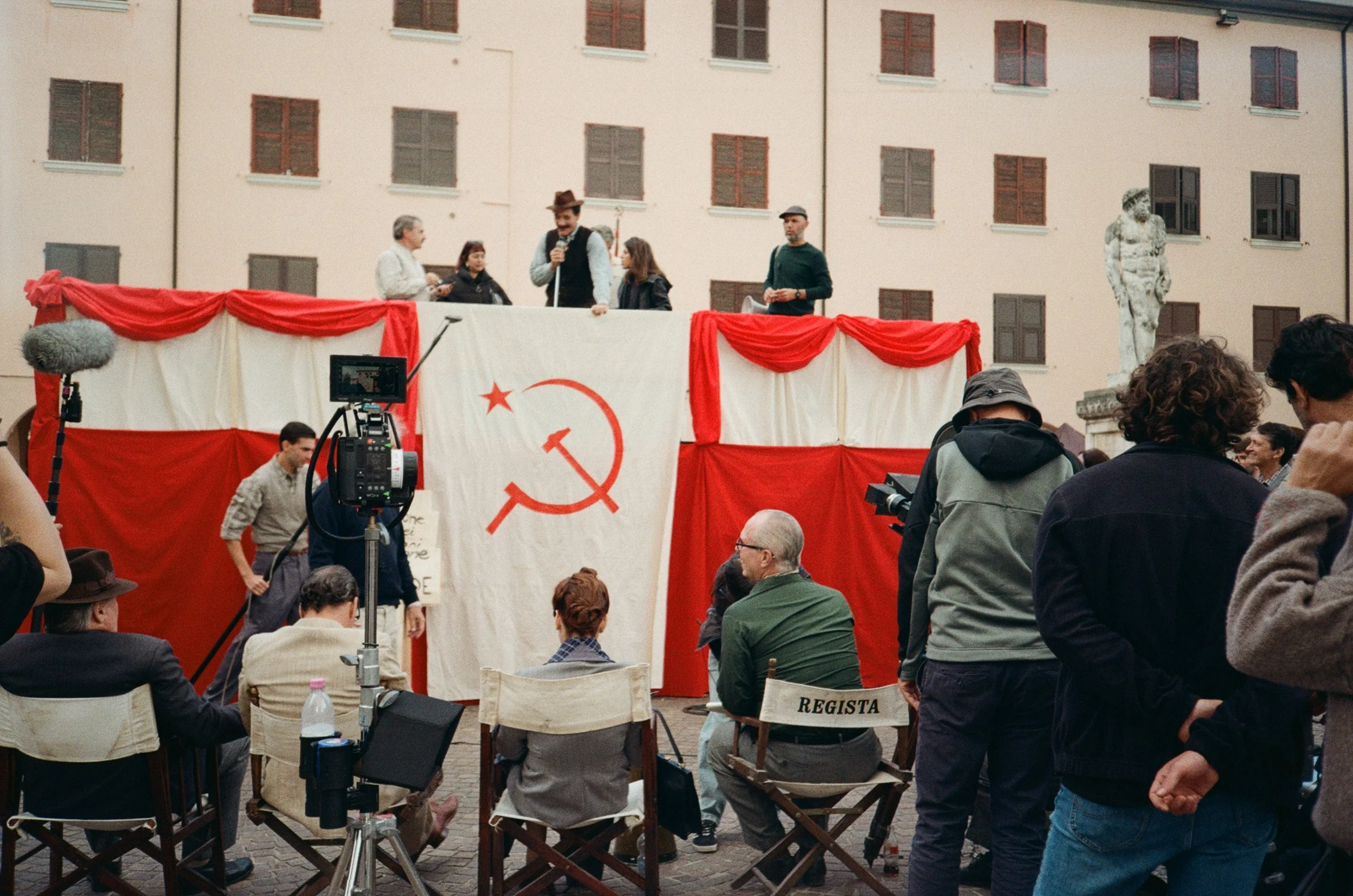 Persone che assistono a uno spettacolo di teatro o una manifestazione, con uno sfondo di drappi rossi e bianchi e il simbolo comunista, una falce e martello, visibile alla scena. Ci sono anche attrezzature per riprese e un pubblico seduto e in piedi.
