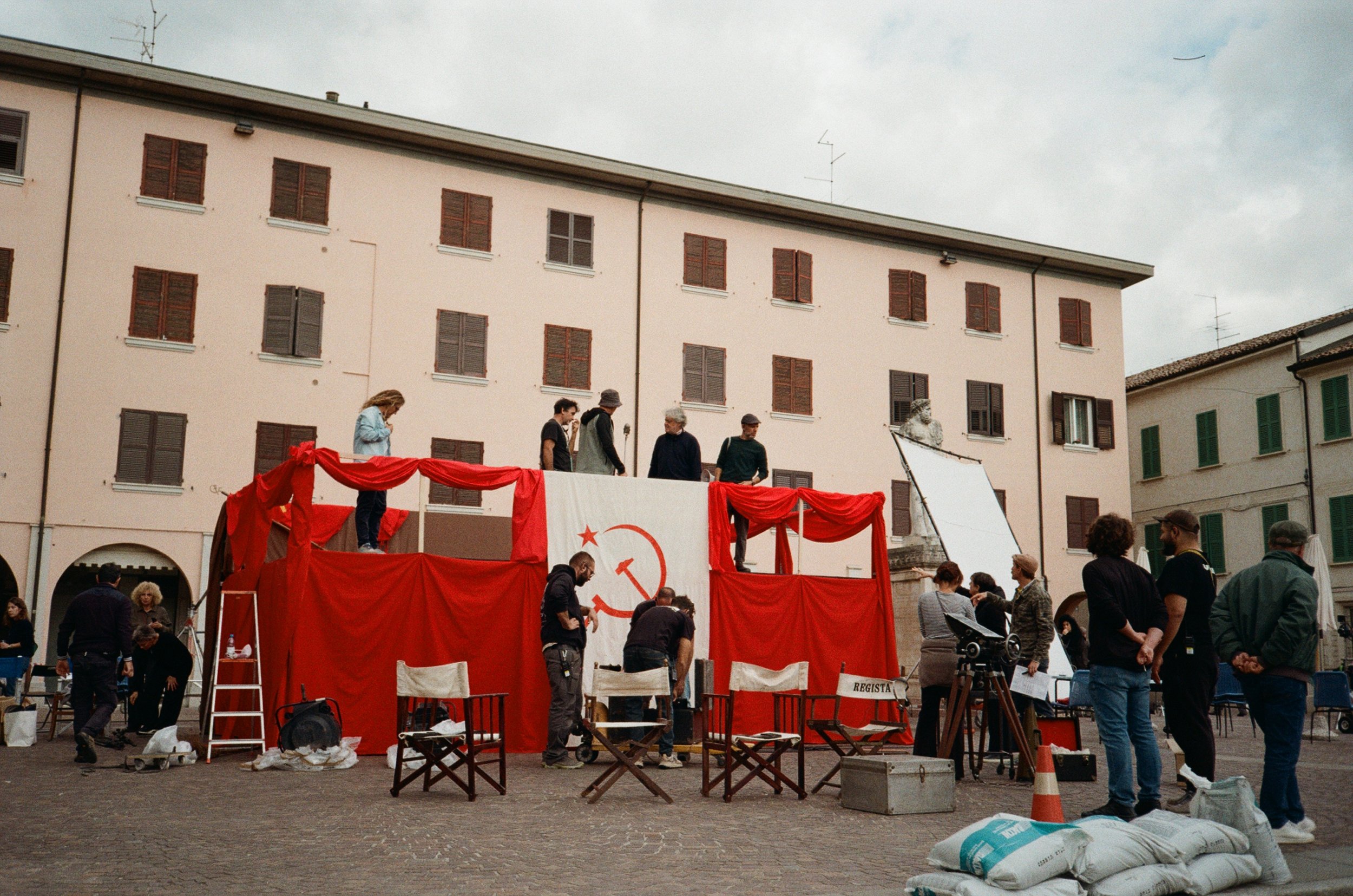 Scena di una produzione teatrale o di un film in un luogo pubblico, con attrezzature e persone impegnate nel set, decorazioni rosse, e un grande pannello con il simbolo del comunismo.