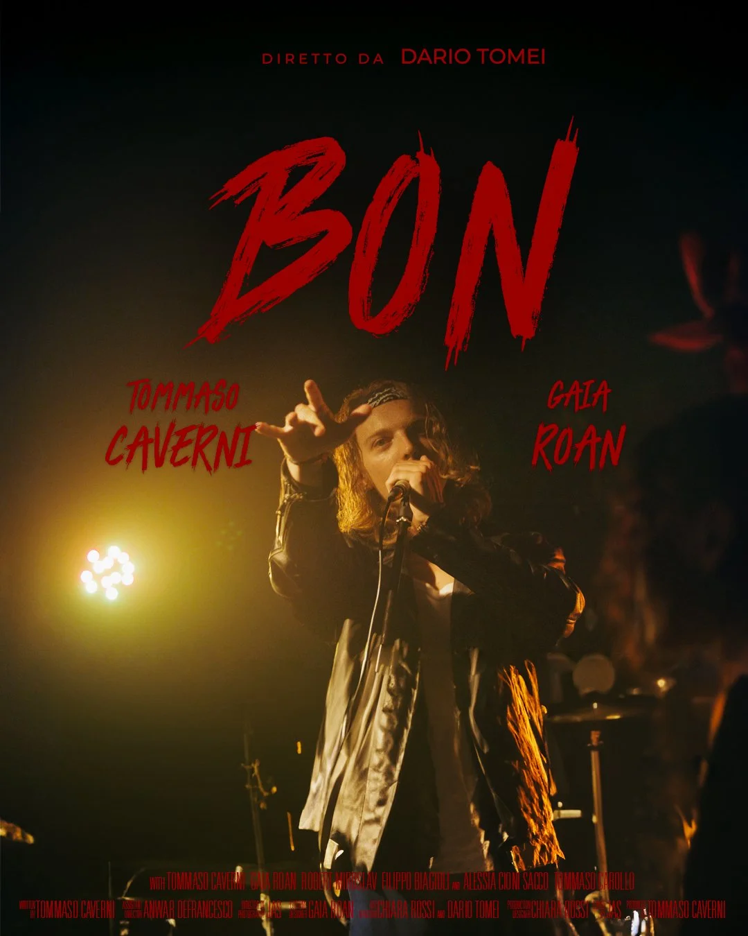 Locandina teatrale con un'immagine di un cantante, Jon Bon Jovi, interpretato da Tommaso Caverni che canta con un microfono, sfondo scuro, titolo Bon scritto in rosso con stile graffito e altri nomi degli attori.