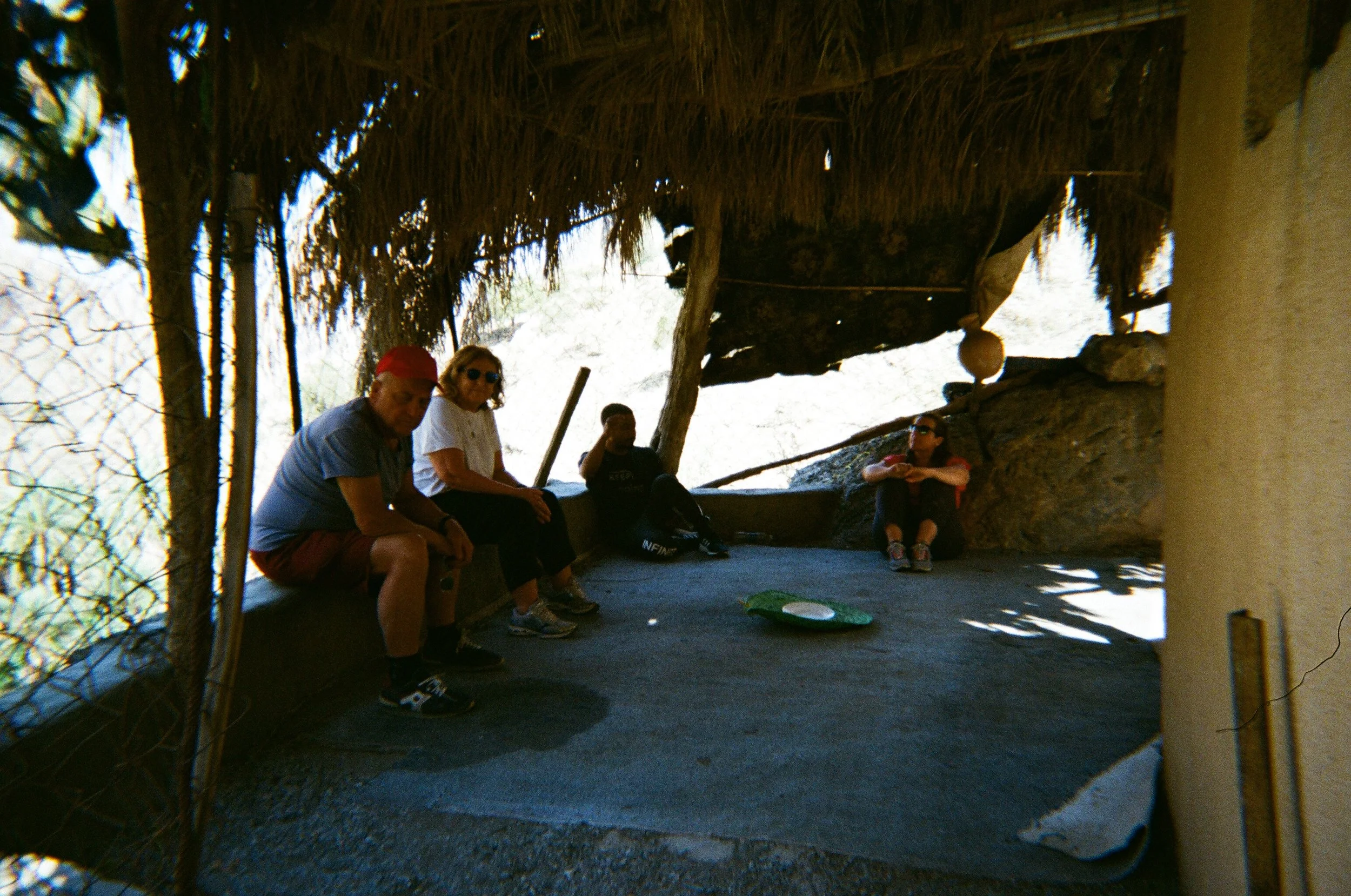 Gruppo di persone che riposa in un rifugio isolato con tetto di paglia, seduti su una piattaforma di pietra, con un fondo naturale illuminato dalla luce.