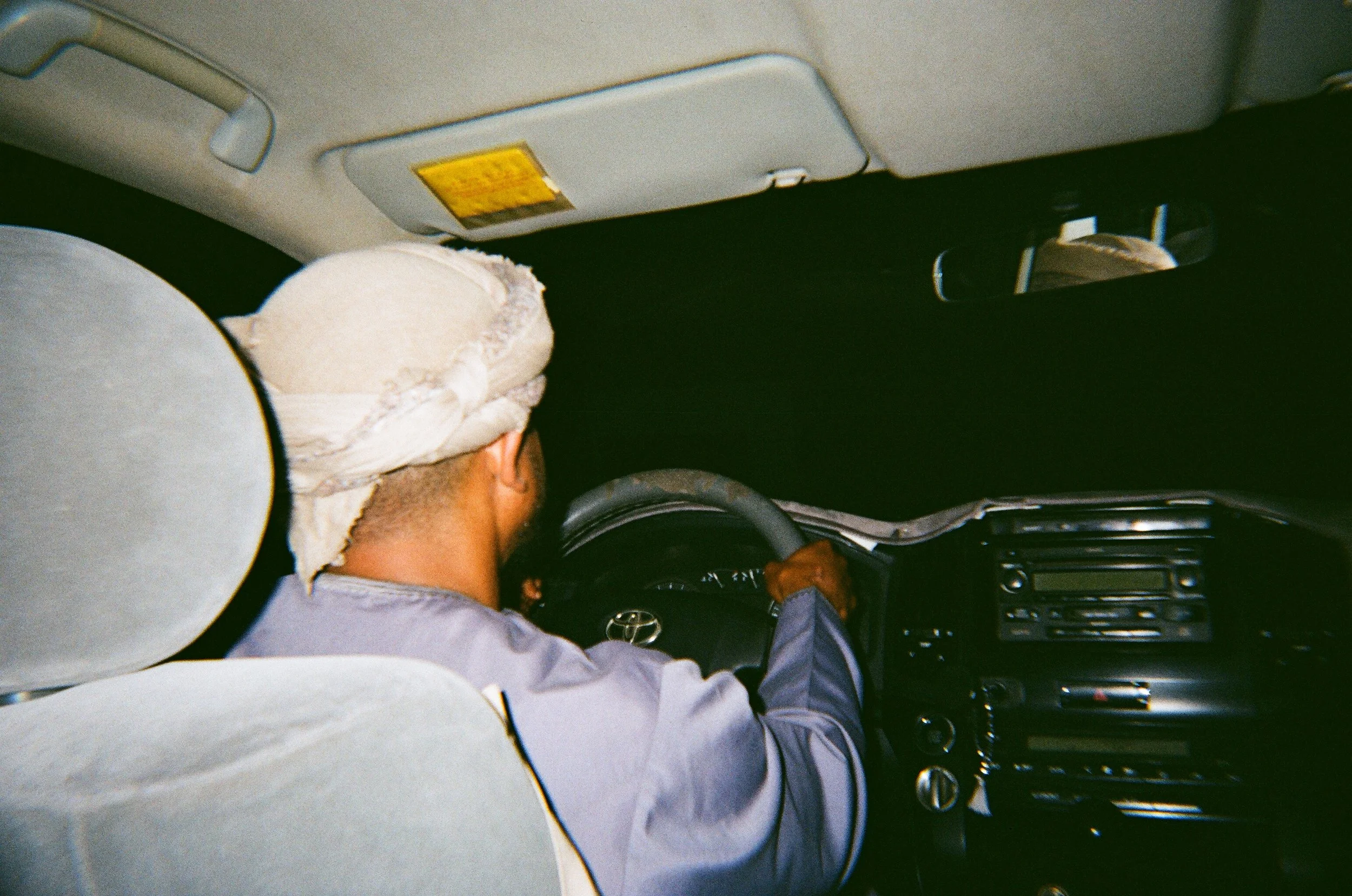 Uomo che guida un'auto, indossa un turbante bianco, visto di spalle, all'interno di un veicolo al buio.