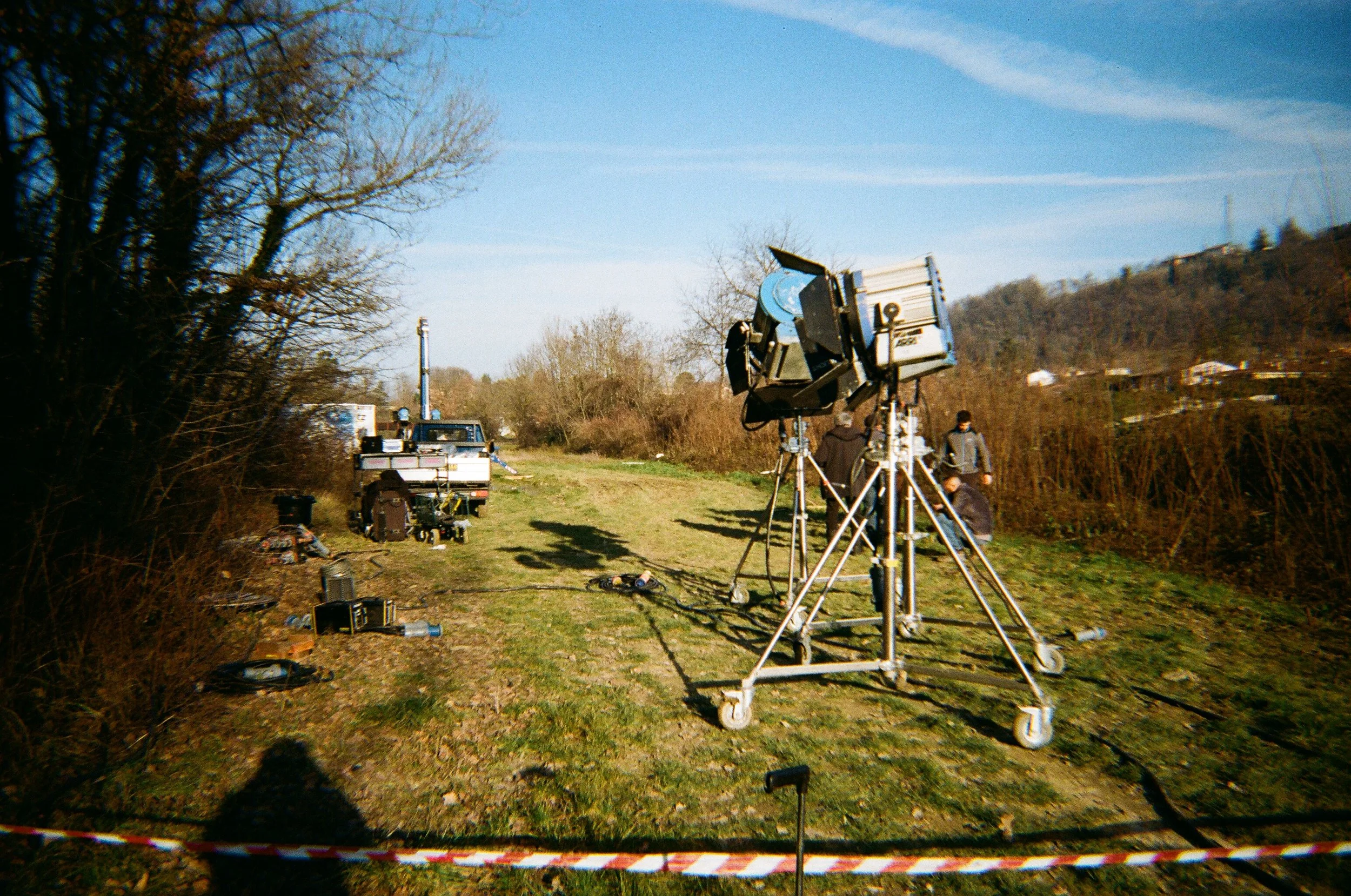 Scena di ripresa cinematografica all'aperto con illuminatori e attrezzature di ripresa su un campo erboso, con quattro persone che si preparano e un cielo azzurro.